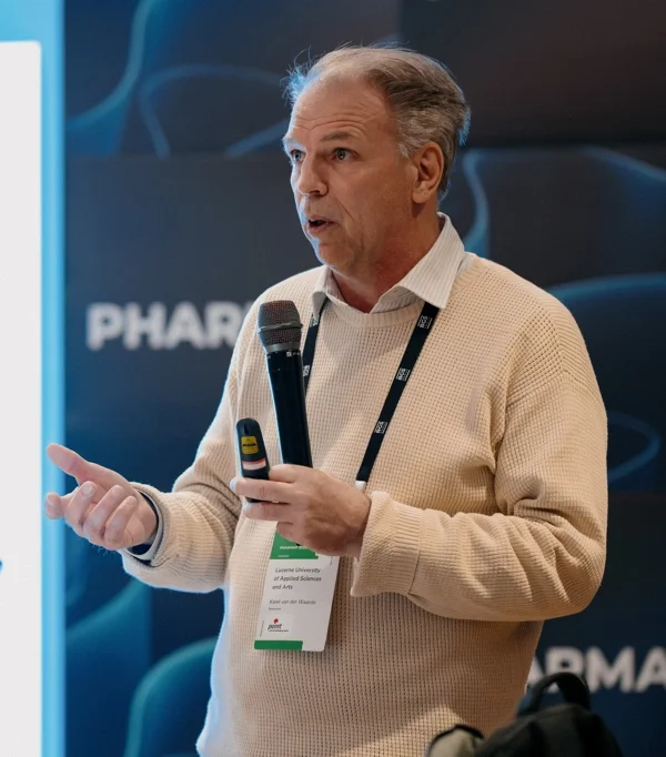 A man standing speaking into a microphone in front of a blue screen with the word "pharma" on it. He is a white-skinned male with gray hair, mouth open mid-speech. He wears a beige sweater with a white collar and a tag on a lanyard around his neck.