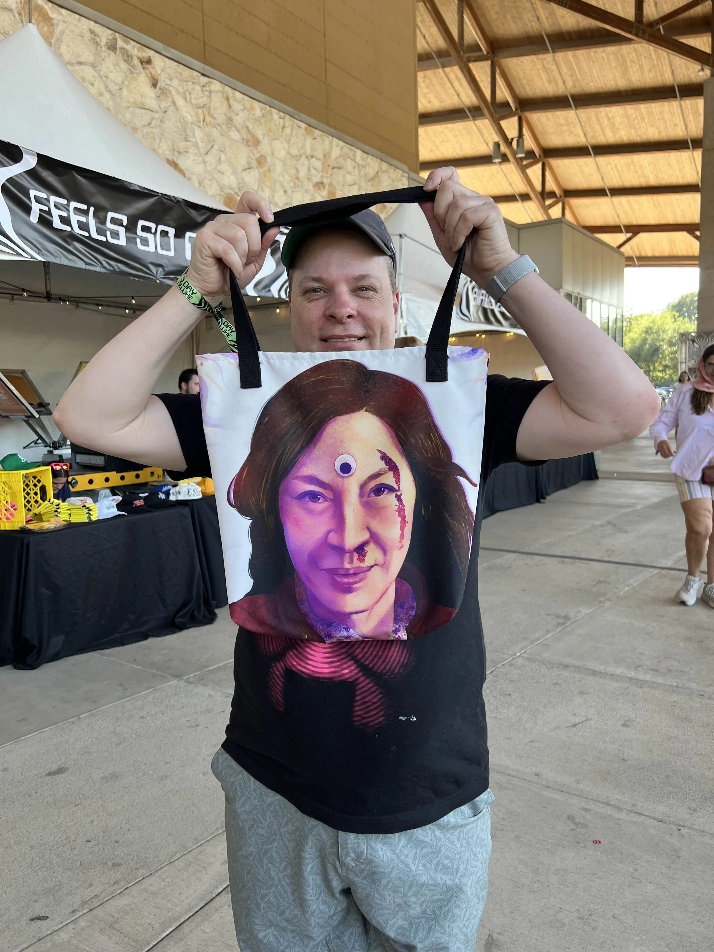 Man stands outside and holds the straps of a bag over his head. Bag has Michelle Yeoh from Everything Everywhere All At Once. Merch booth behind him and there's a concrete floor. He wears black hat, black T-shirt, gray shorts, green strap on wrist.