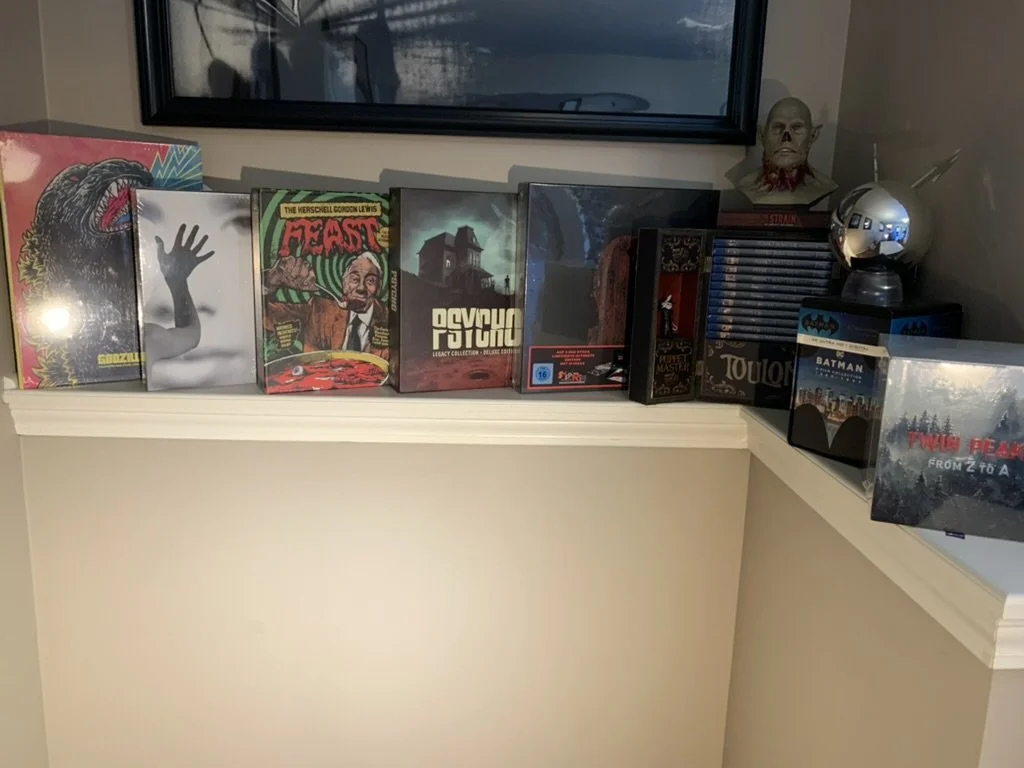 A shelf full of Blu-ray boxsets on a beige wall. From left to right is Godzilla, Ingmar Bergmanm, Herschell Gordon Lewis, Psycho, Batman, Twin Peaks, and several less visible sets. A monster head replica sits on one boxset, along with a Phantasm orb.