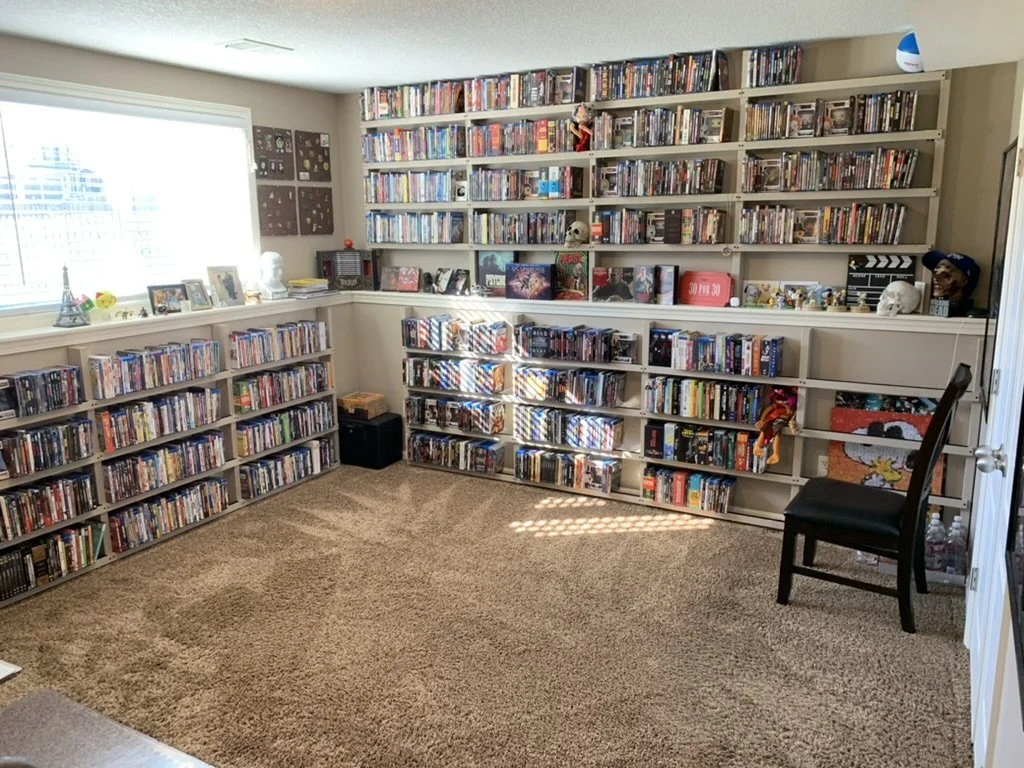 A carpeted room with beige walls and a window bright from sun has walls with built-in shelves neatly lined with Blu-ray movie cases, plus assorted other memorabilia, including skulls and a Snoopy print. Black chair in front of a white door on right.