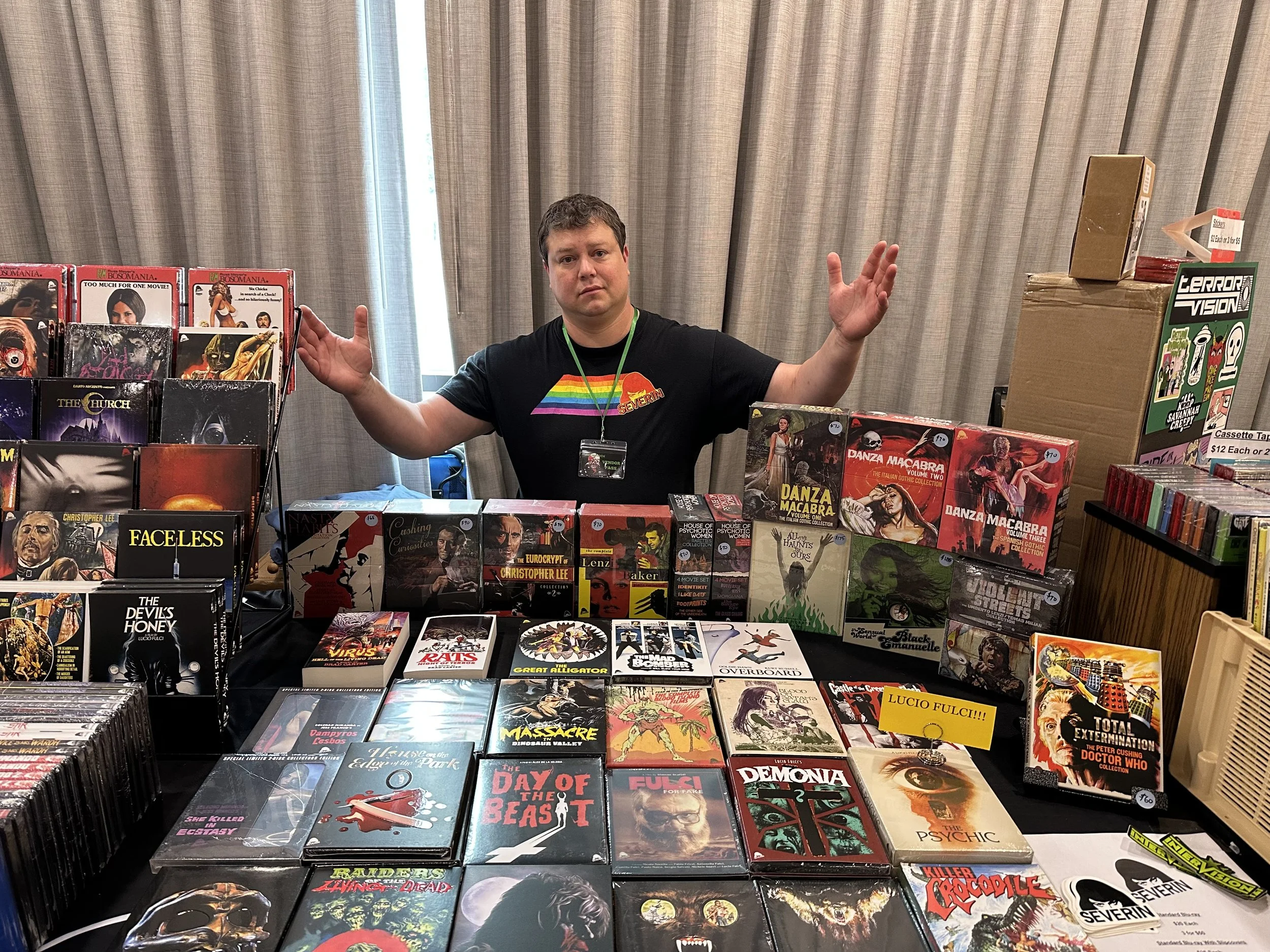 Will sits at booth covered with Blu-ray movie cases and box sets in front of a gray curtain. His hands are extended ahead of him. He's wearing a black T-shirt with a rainbow version of the Severin logo, which is company name with woman's head above.
