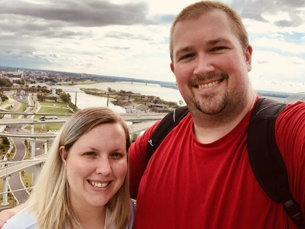 A much shorter woman with blonde hair and white skin stands beside a taller man wearing a red shirt with backpack straps visible. He has a goatee beard and short blond hair. They are standing outside with highways behind them and river-like water.