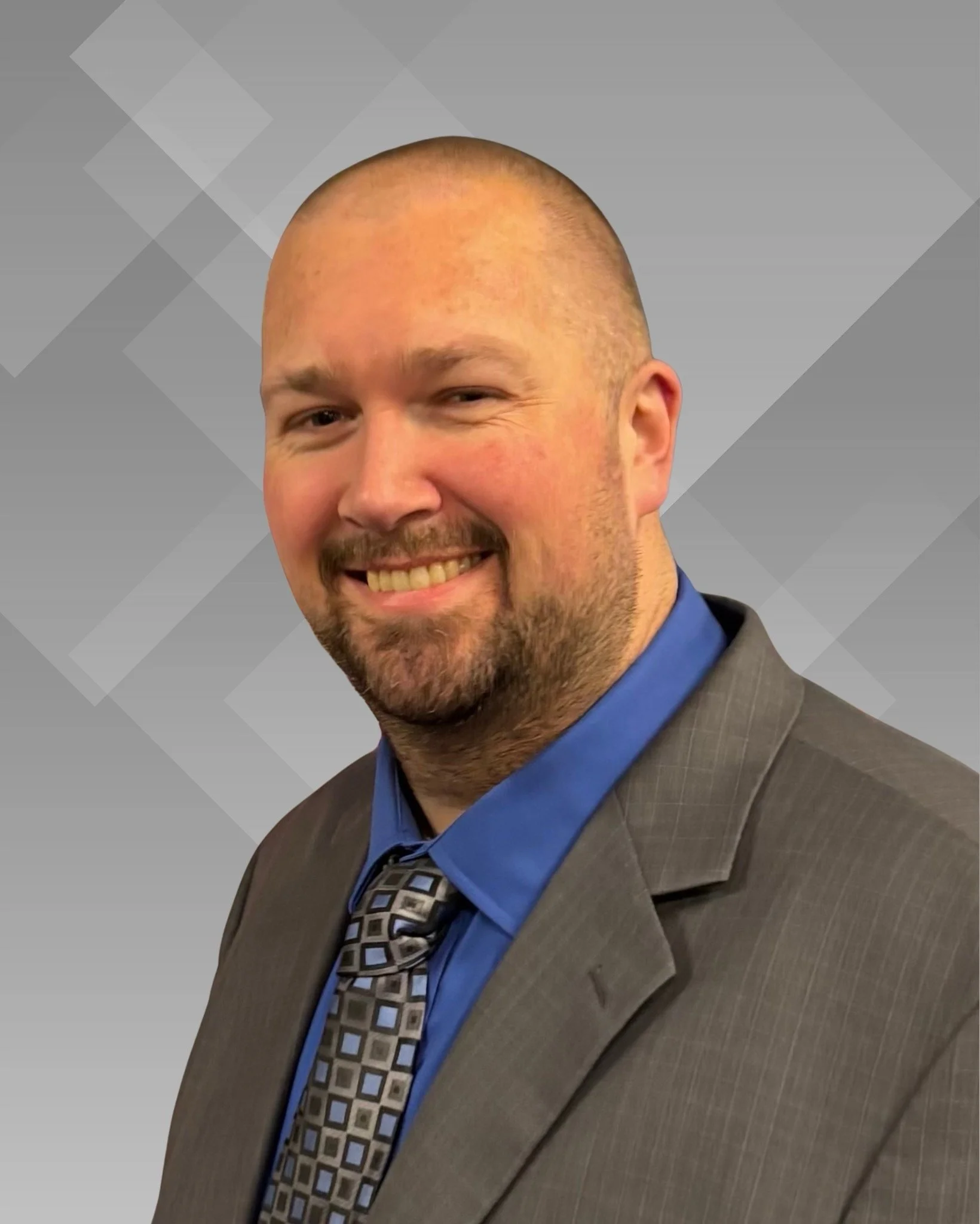A headshot of a man with rosy cheeks,  a shaved head and a short goatee who is smiling. He wears a gray suit that has light small lines with a blue dress shirt and a blue/gray chequred tie underneath. Nearly solid silver background.