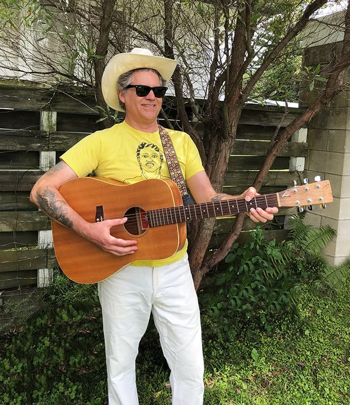 A man stands outside holding acoustic guitar. Wears white cowboy hat, sunglasses, yellow T-shirt with animation of person, and white pants. Wooden fence (his height) behind him. Unkempt lawn and bushes, and scraggly tree with many small branches.