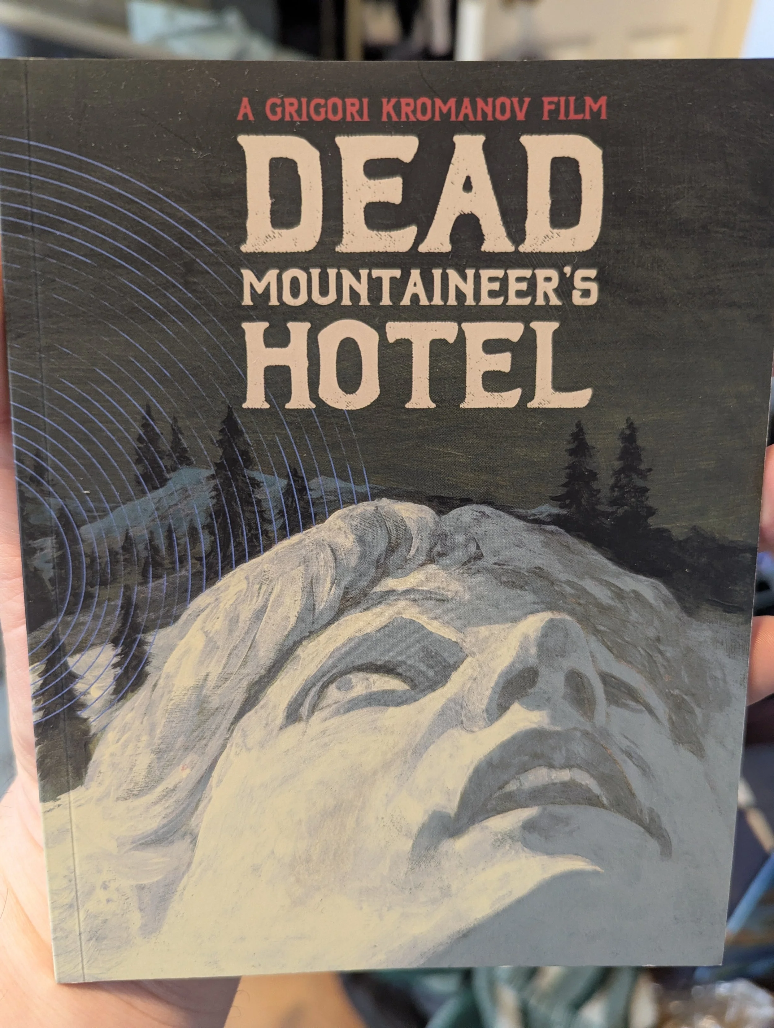 Front cover of Dead Mountaineer's Hotel booklet, which has film title at top over a night image of a mountain in winter. It's covered with snow. That snow has the image of a person's face in it, looking up with mouth open and worried look in eye.
