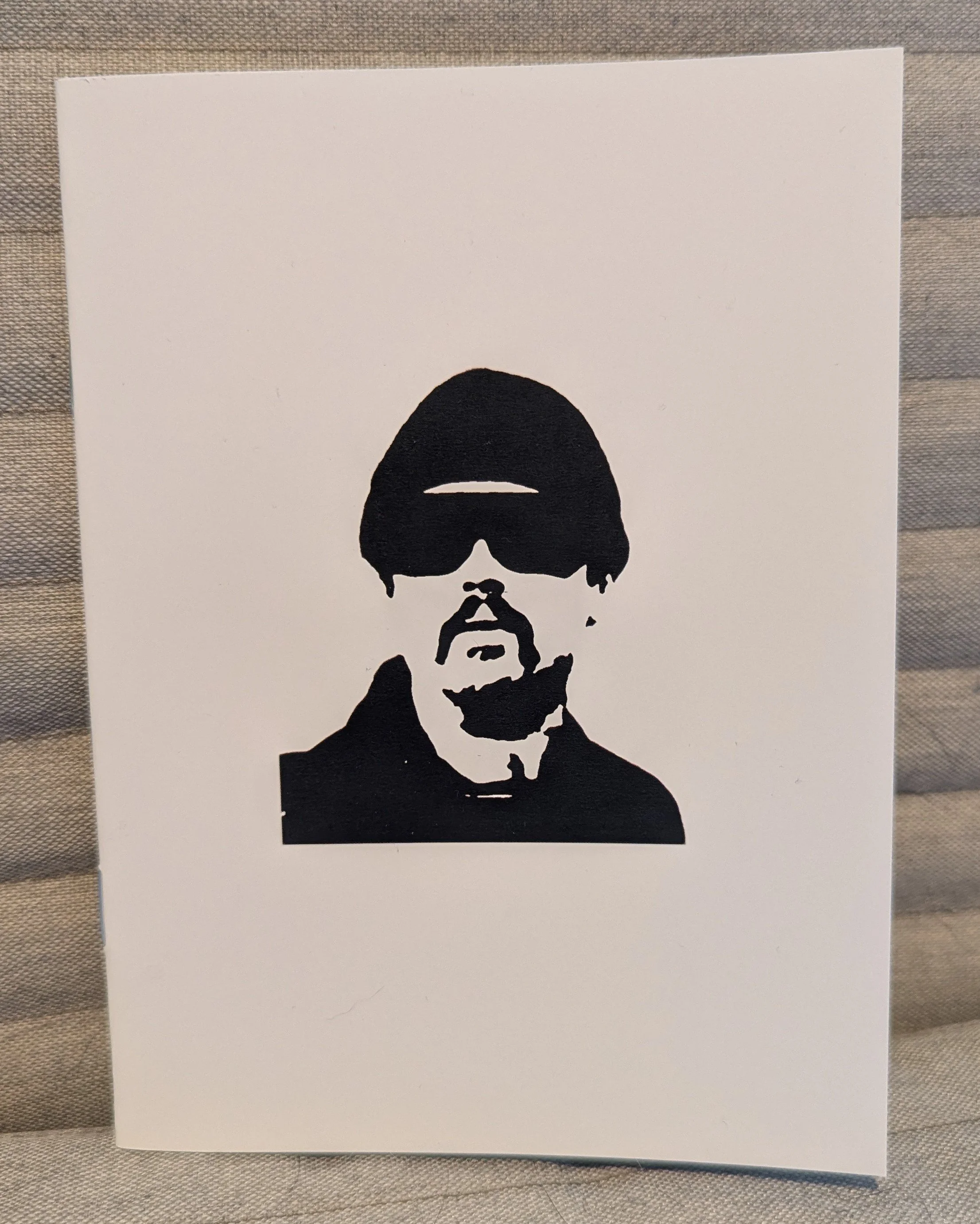 Close up of the booklet coer, which has a sketch of a man (Leonardo DiCaprio's character in the film) in ink black on an all-white background. Behind booklet is tan fabric. The character has big black shades, hat, goatee, shadow on neck, black shirt.