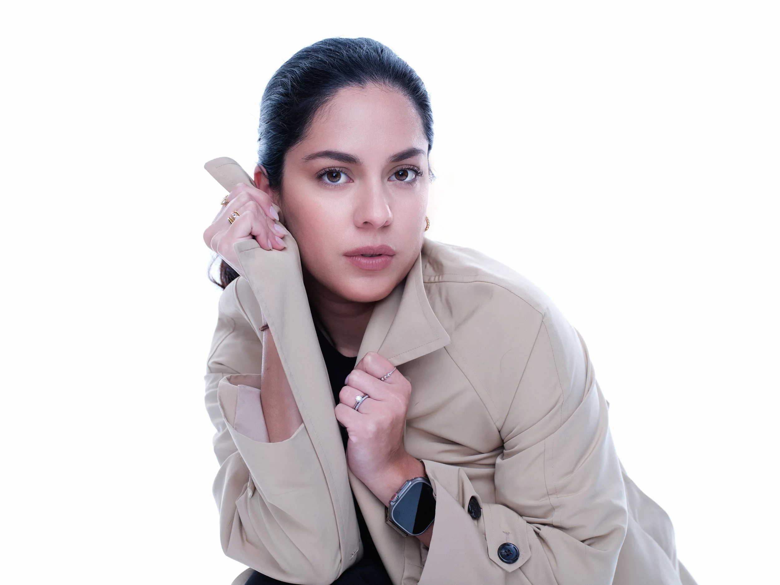 A woman with dark hair, wearing a beige trench coat and a smartwatch, holds a vintage telephone receiver to her ear against a white background.