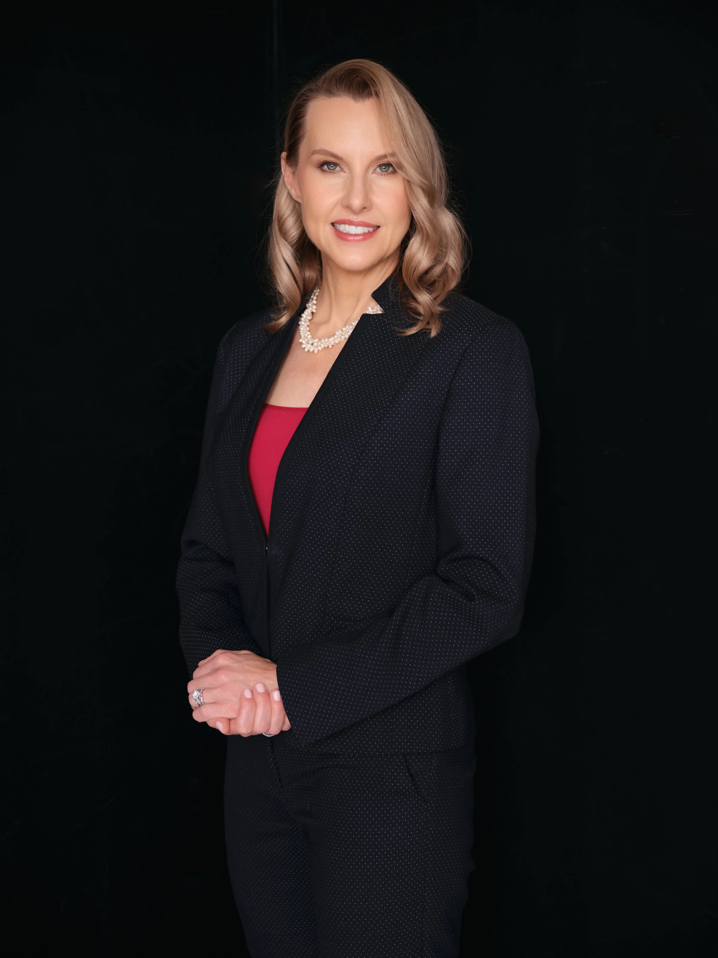 A woman with blonde hair styled in loose waves, wearing a dark blazer with small white polka dots over a red top, accessorized with a pearl necklace and ring, standing against a black background and smiling at the camera.