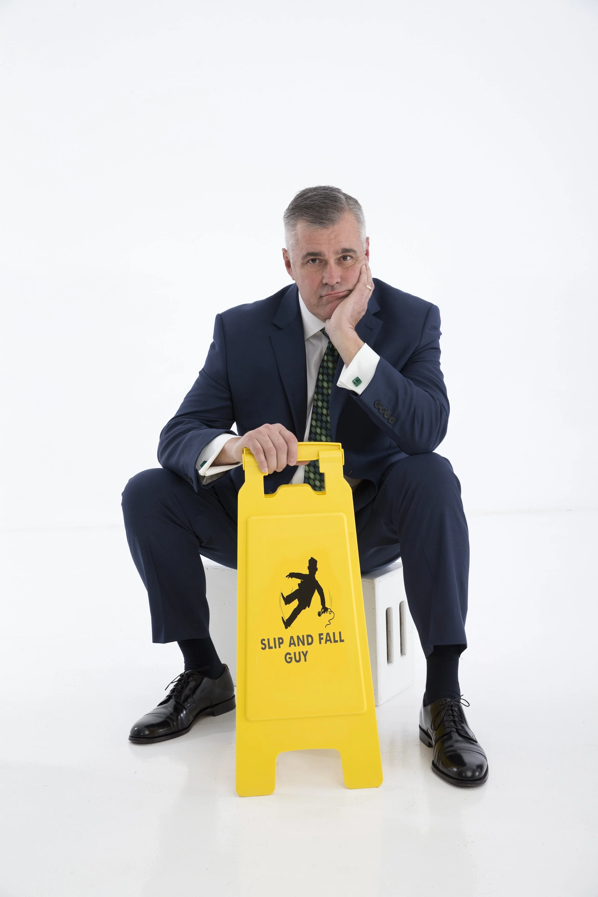 A man in a navy suit sitting on a white stool with a yellow slip and fall sign that says 'Slip and Fall Guy', looking tired or bored, against a plain white background.