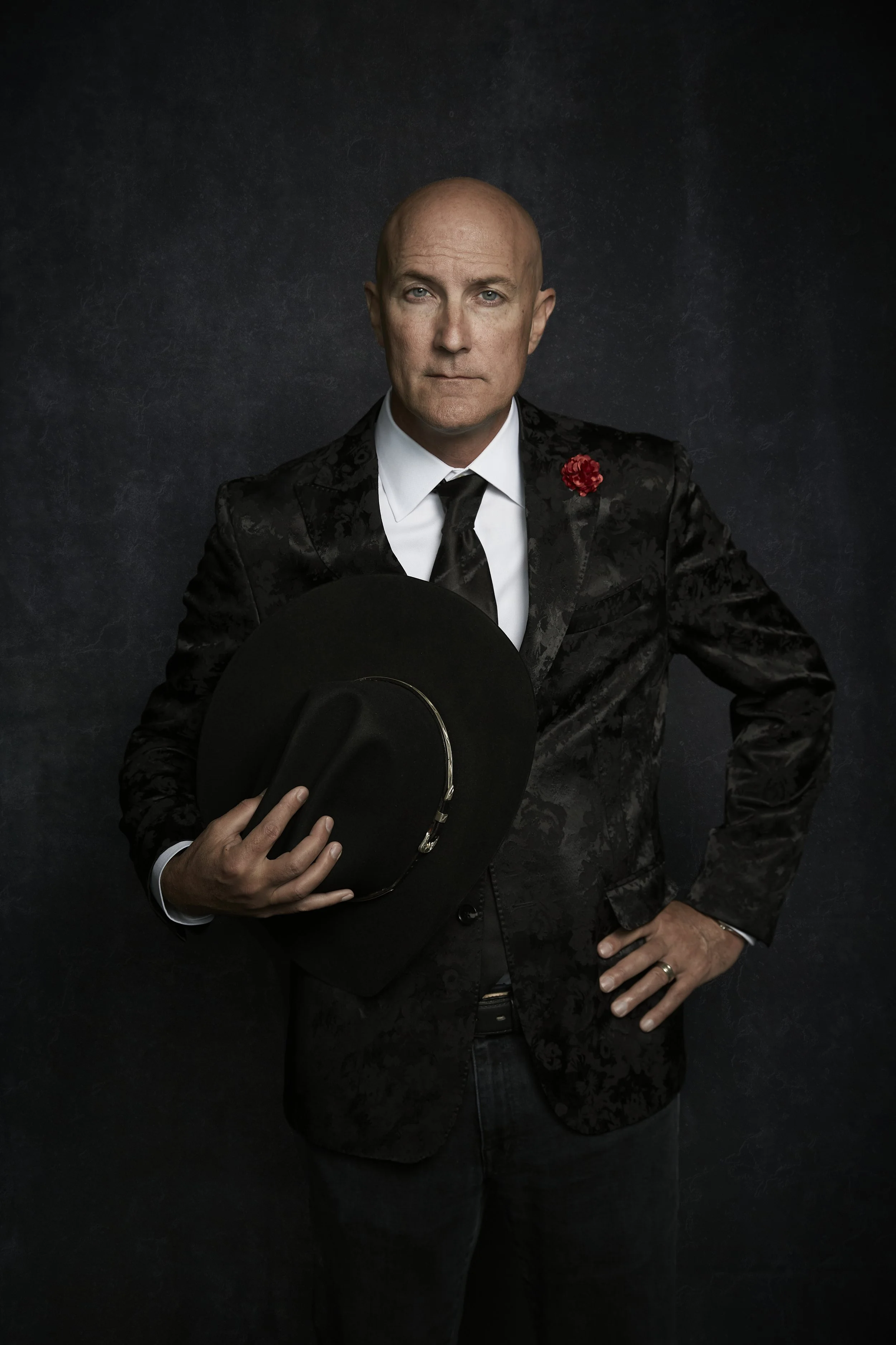A bald man in a black patterned blazer, white shirt, and black tie holding a black hat, standing against a dark textured background.
