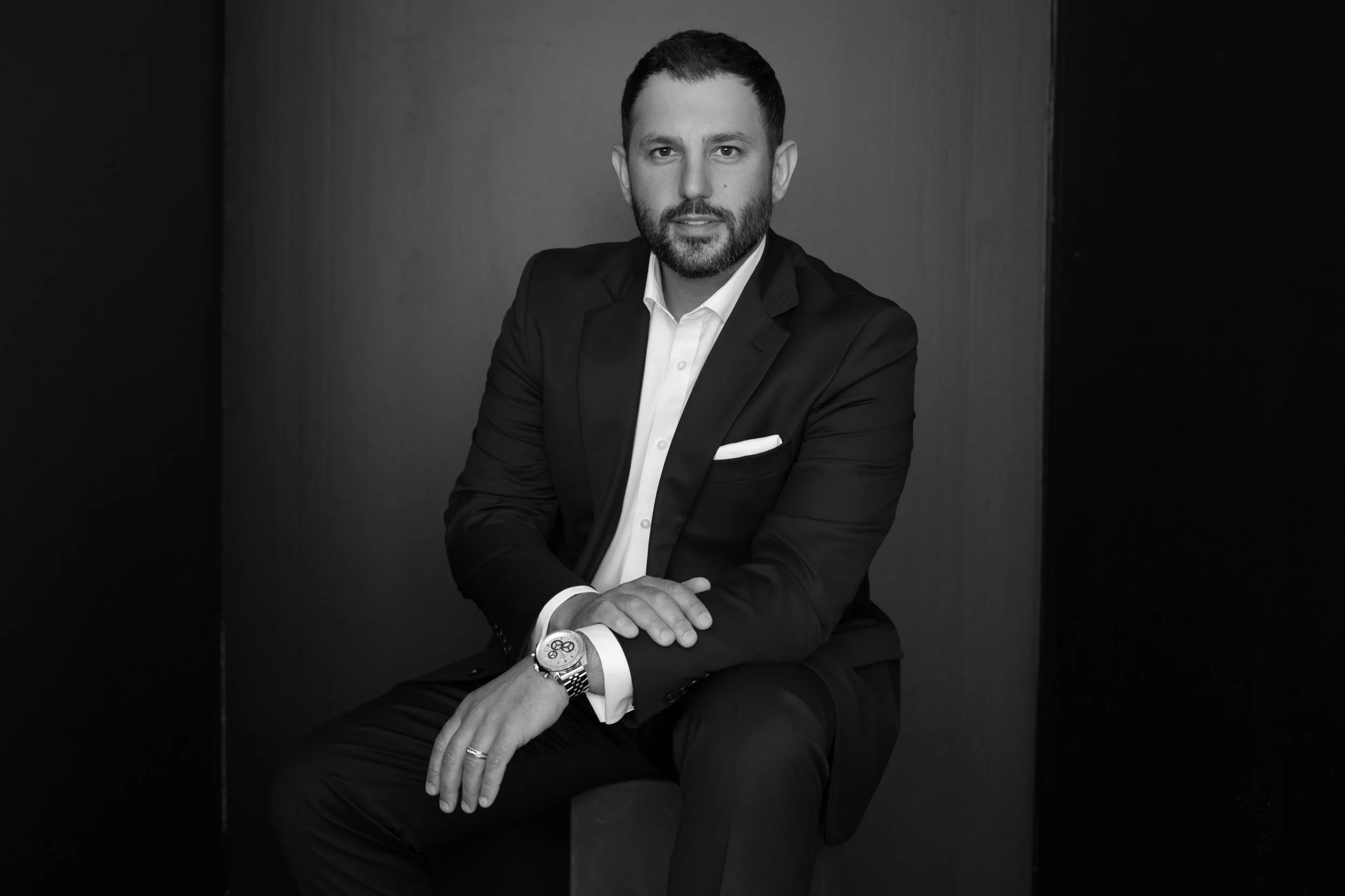 A black and white portrait of a man in a suit and white shirt, sitting with his hands clasped and looking at the camera.