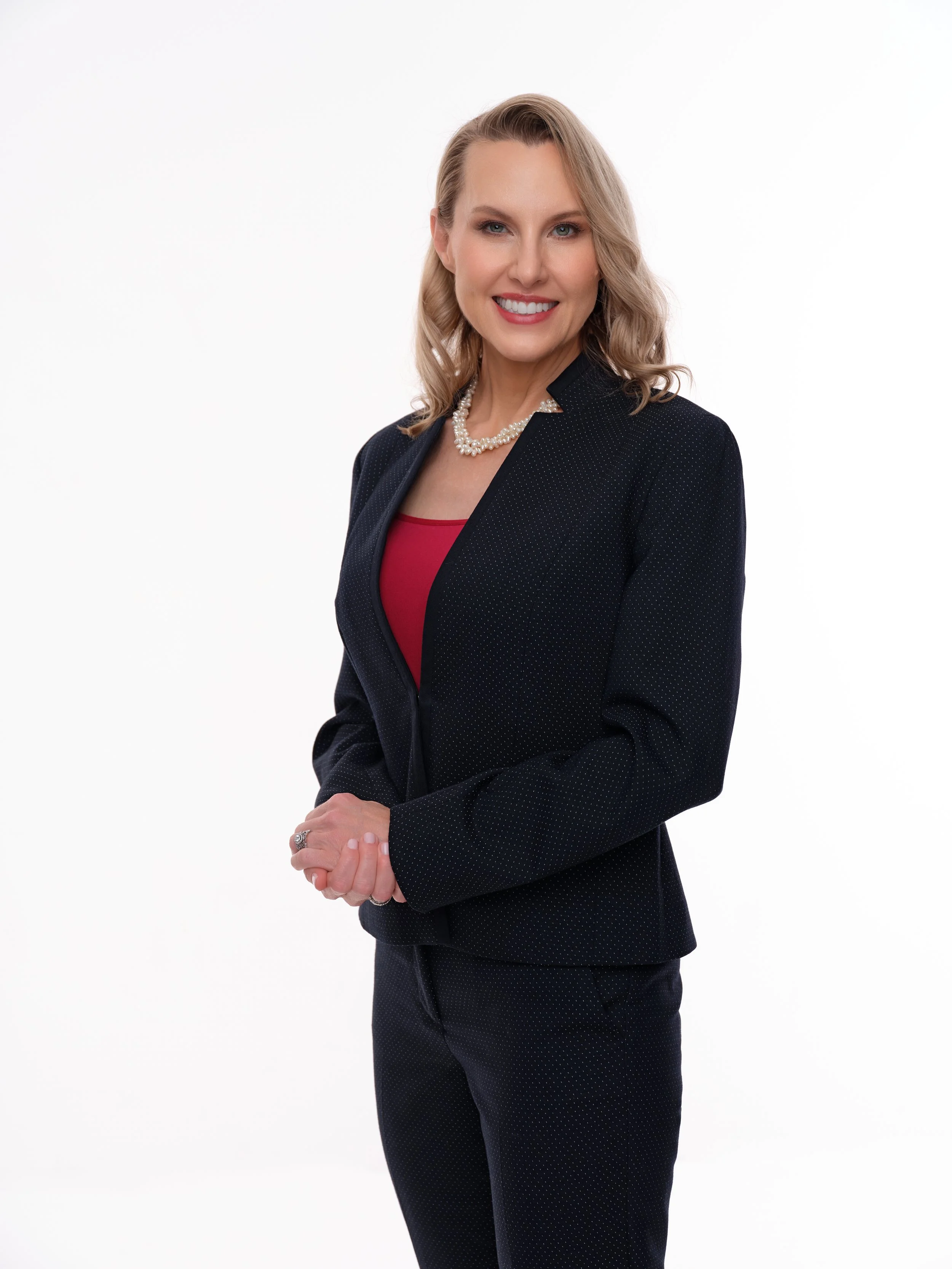 A professional woman with blonde hair, smiling, wearing a dark blazer over a red top, with a pearl necklace, posing against a white background.