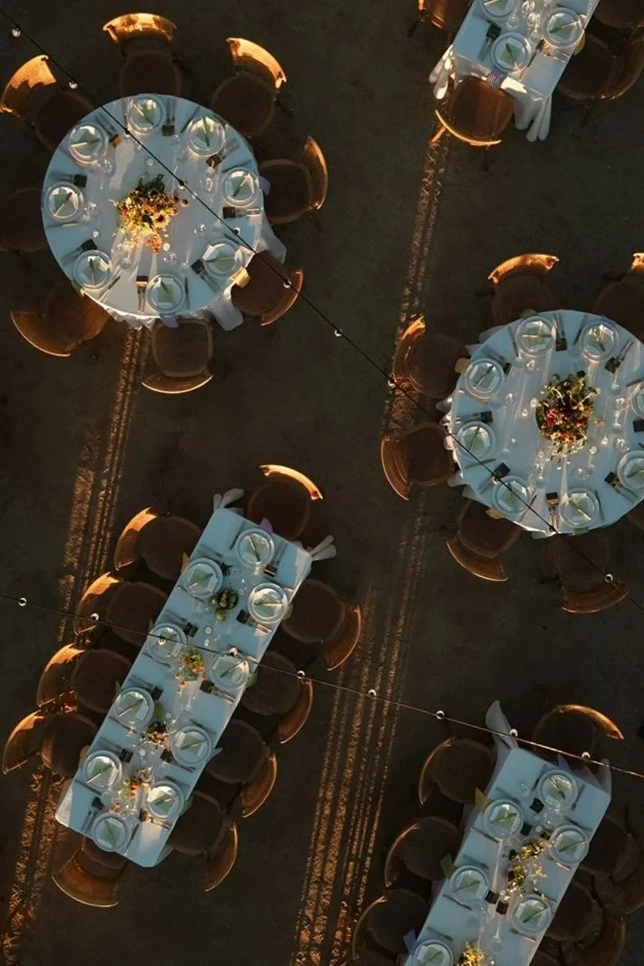 Overhead view of four round tables and two rectangular tables set for a formal event, with white tablecloths, glassware, plates, and floral centerpieces, illuminated by warm lighting.