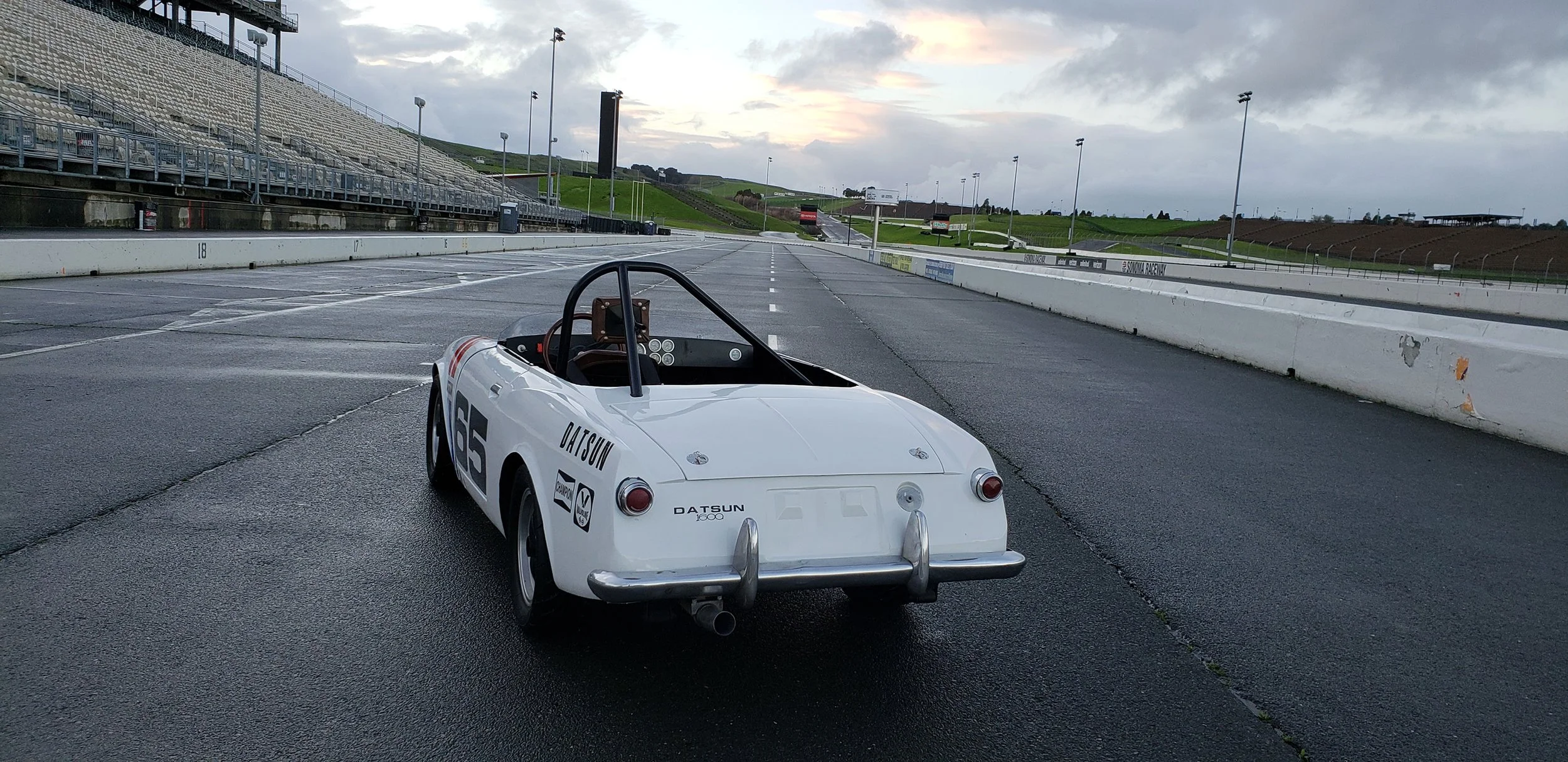 Rear angle view photo of a vintage 1968 white race livery Datsun 1600 Roadster Fairlady car in a race track location. Bespoke build by premium car restoration and modification workshop Retro Sporting in California.