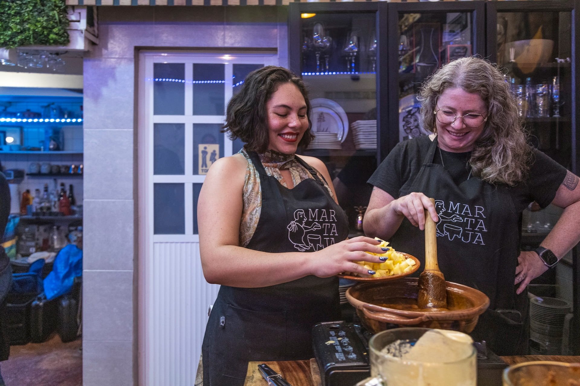 A smiling guest of Martaja adds diced pineapple and apple to a handmade clay pot with mole in it in a cooking class.