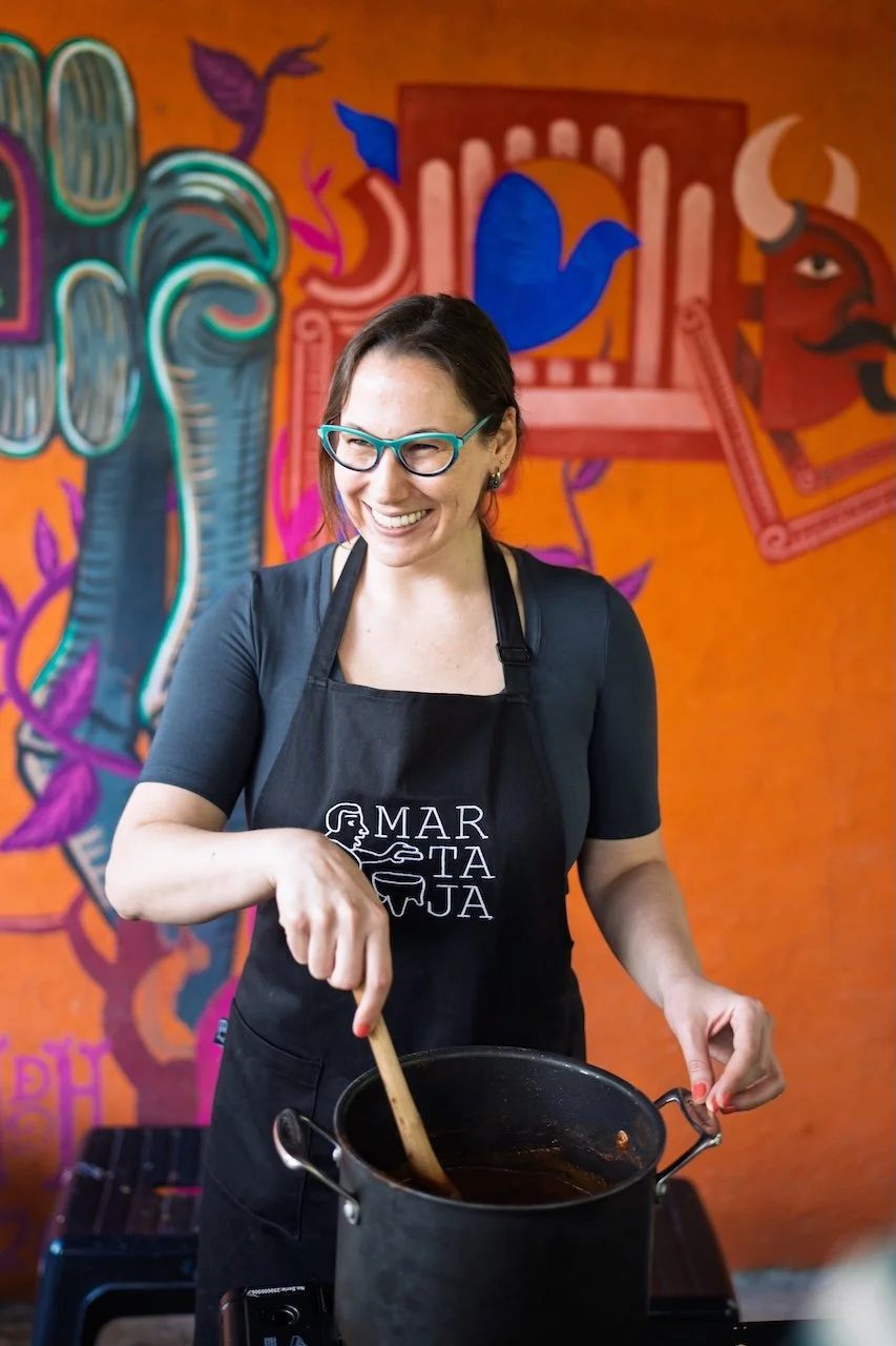 A smiling guest stirs mole during a rooftop cooking class in Condesa, with a colorful mural behind her