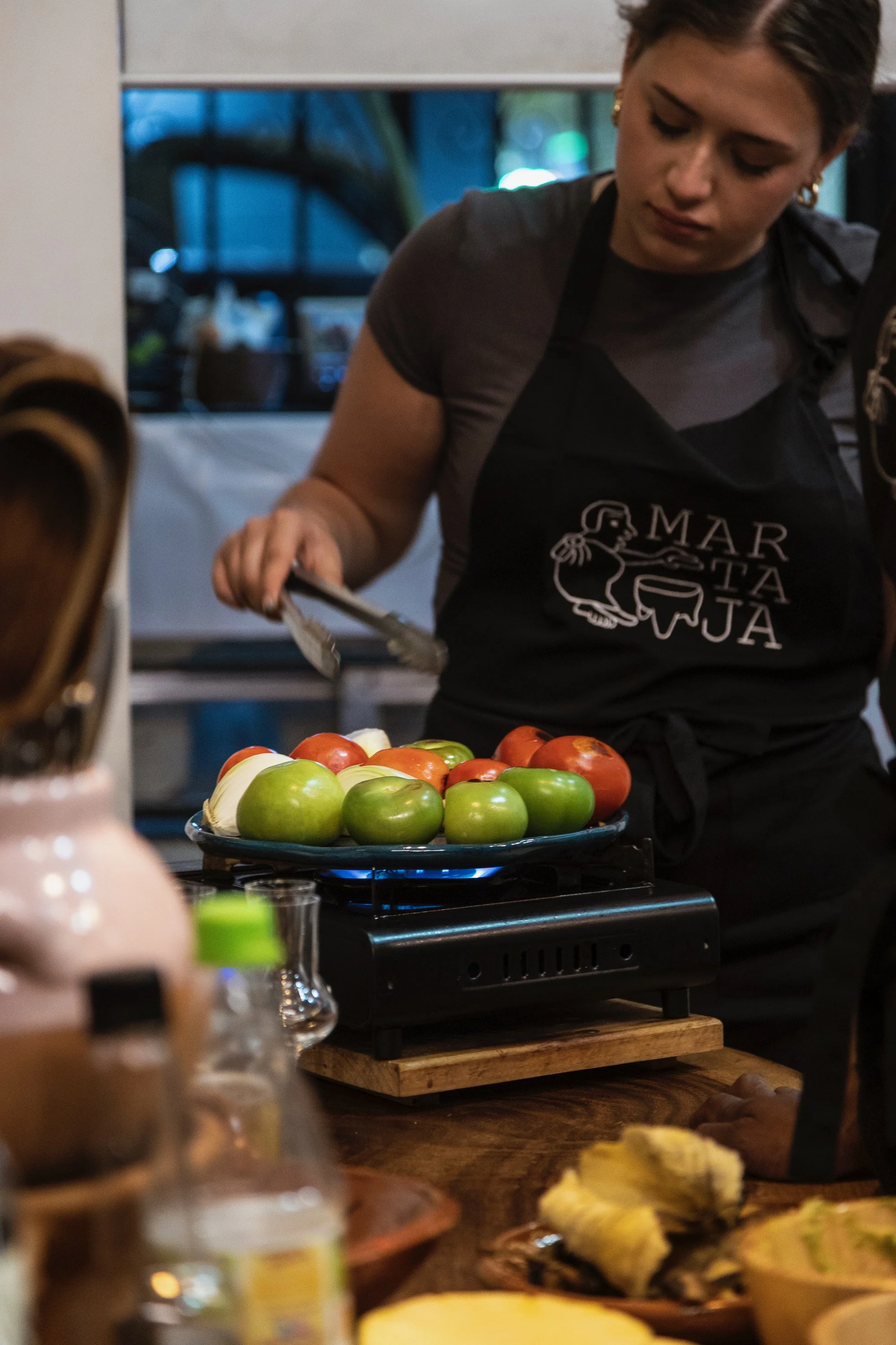 A woman wearing a black apron preparing food at a market or event, with various fruits and dishes displayed in front of her.