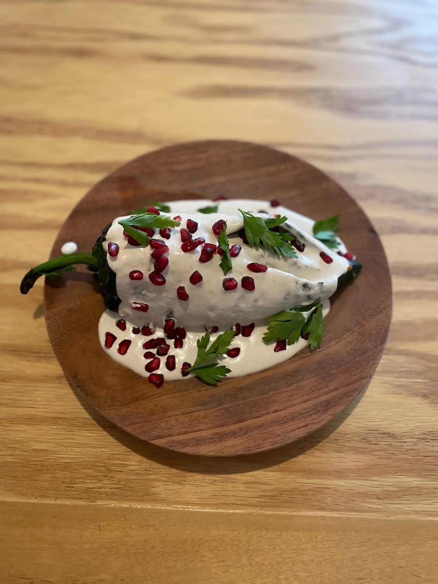 A green poblano pepper covered in creamy white walnut and goats cheese sauce, pomegranate seeds and cut parsley, on a beautiful wooden plate.