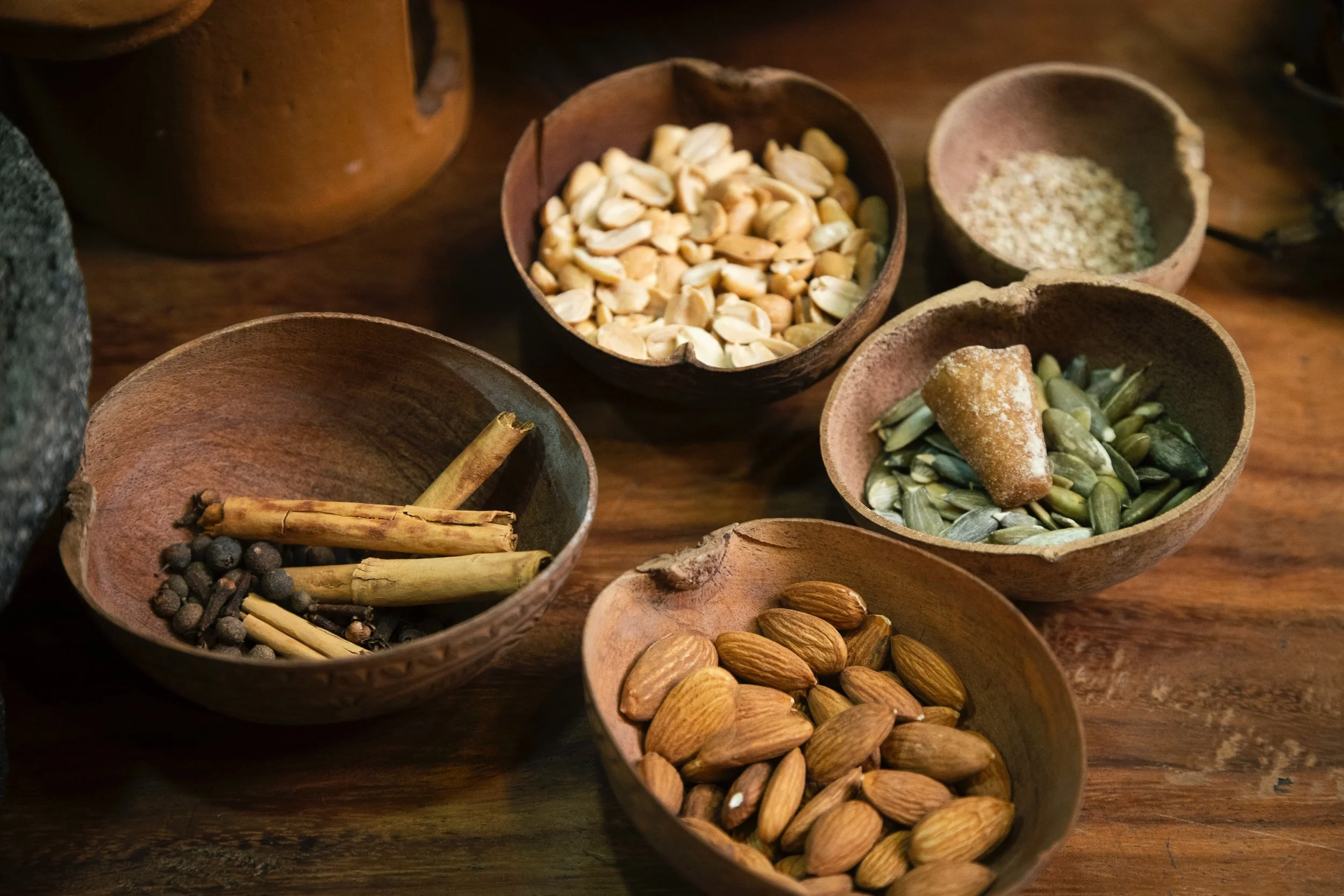 Small wooden bowls filled with various seeds and spices, such as almonds, pumpkin seeds, and cinnamon sticks, arranged on a wooden surface.