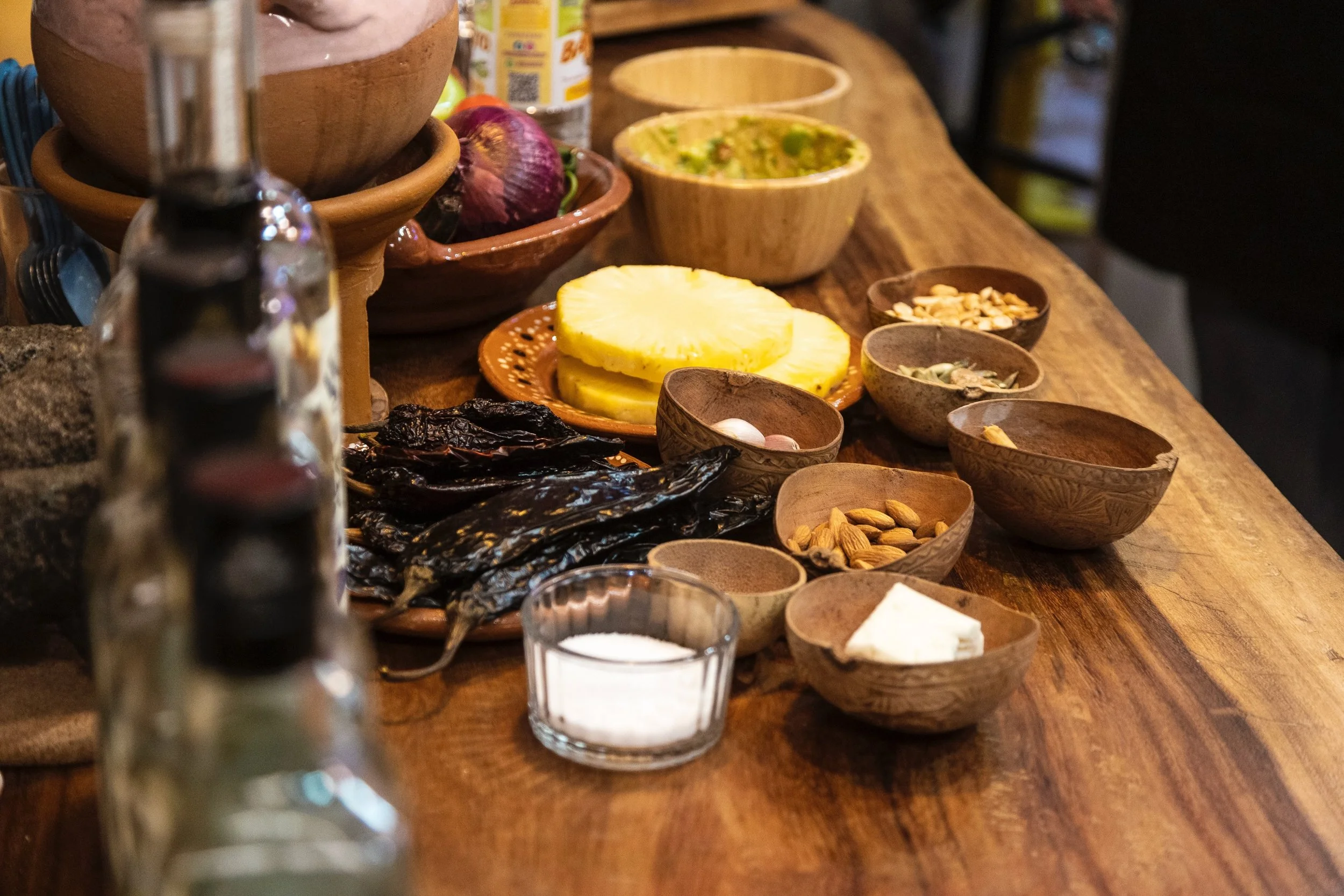 A wooden table with various bowls containing pineapple slices, seeds, and spices, with additional bowls and ingredients in the background.