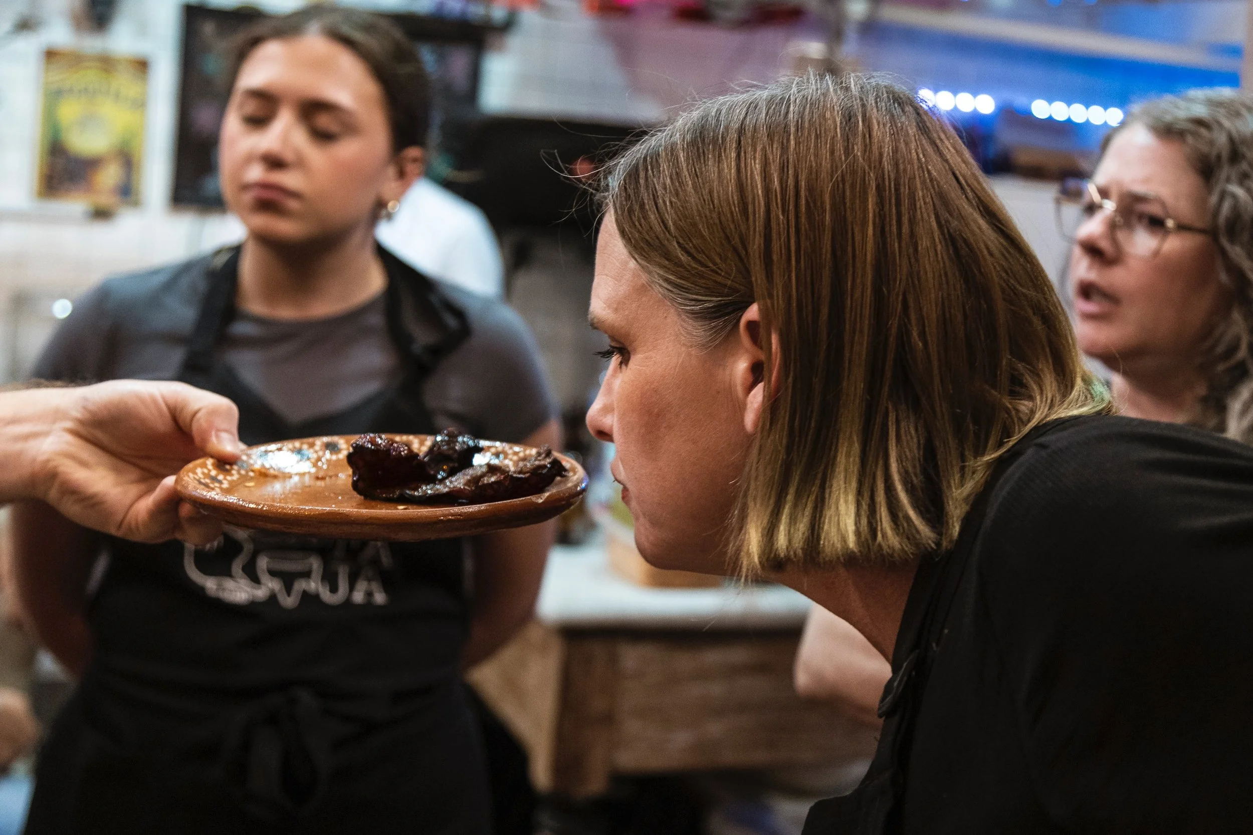 A waitress is holding a plate with food, possibly ribs, as a woman with brown hair looks at it, in a restaurant setting with undistinct background details.