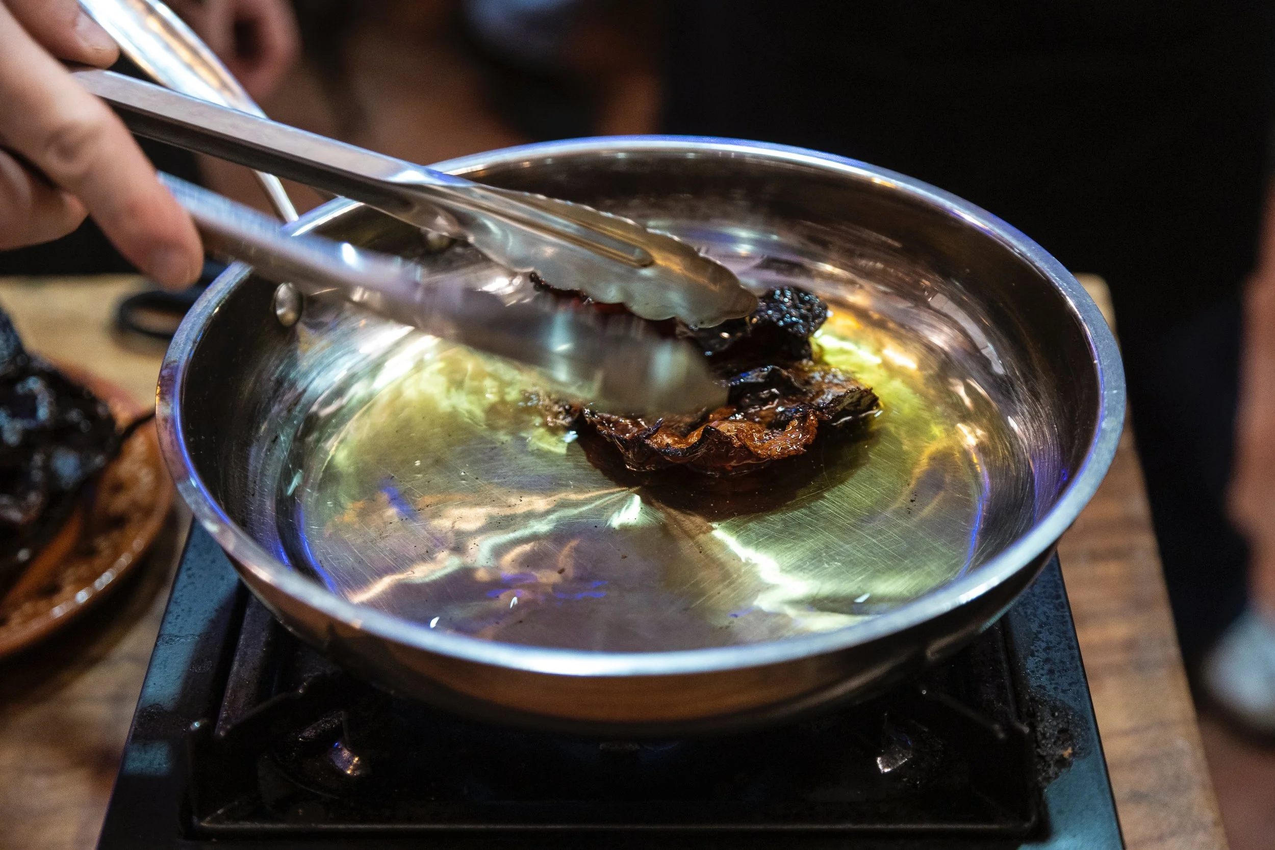 A person is using tongs to hold a piece of food in a stainless steel pan on a stove. The food appears to be dark and caramelized.