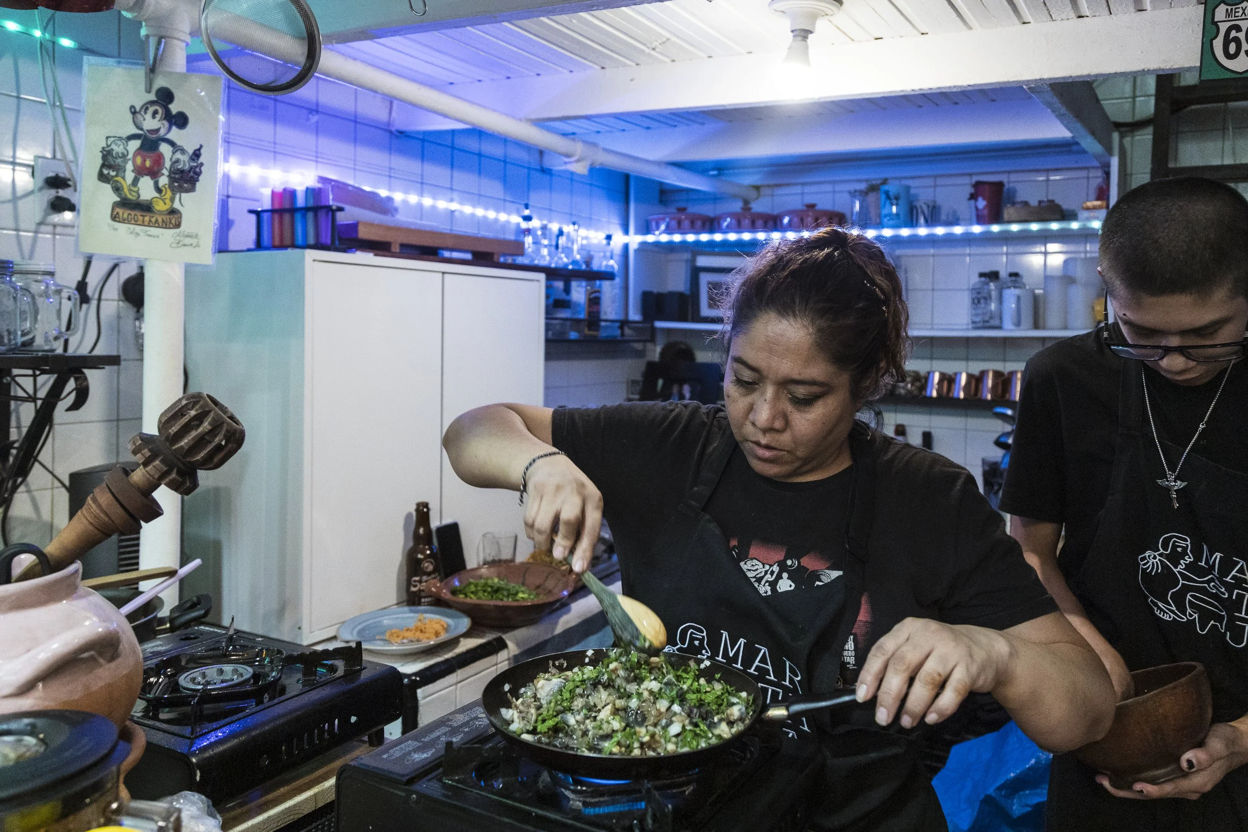 Two people cooking in a kitchen with white tiled walls, one woman and one man, preparing food at a stove. The woman is using a spatula to stir a dish with greens and vegetables in a frying pan.