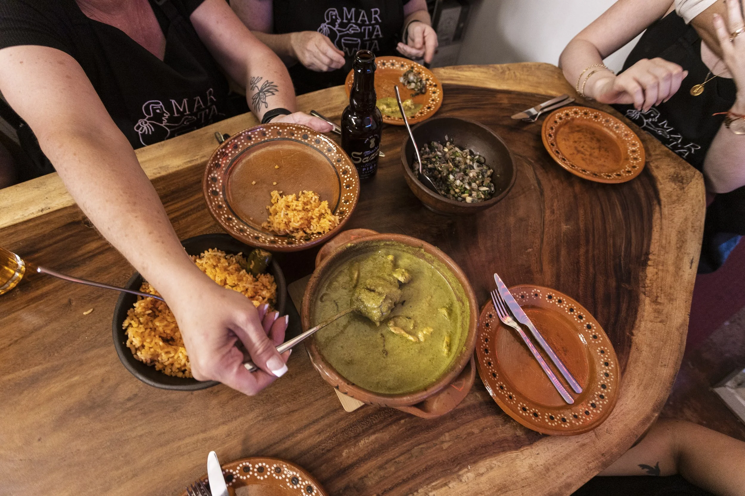 People sitting around a wooden table enjoying Mexican food, including rice, green sauce, and salsa, with several small orange and black patterned plates and a bottle of beer.