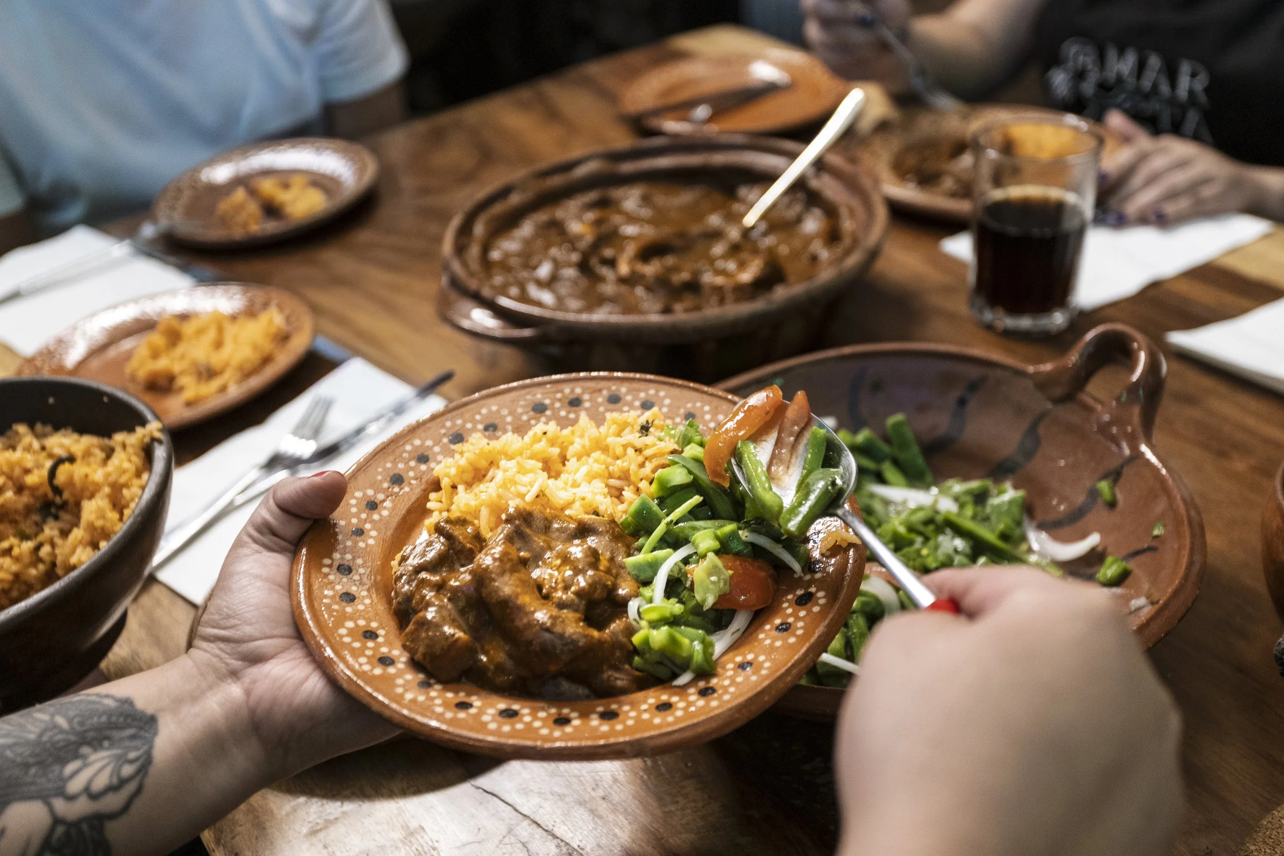 Hand holding a small clay plate with rice, beef stew, and vegetables, at a table with other dishes and drinks.