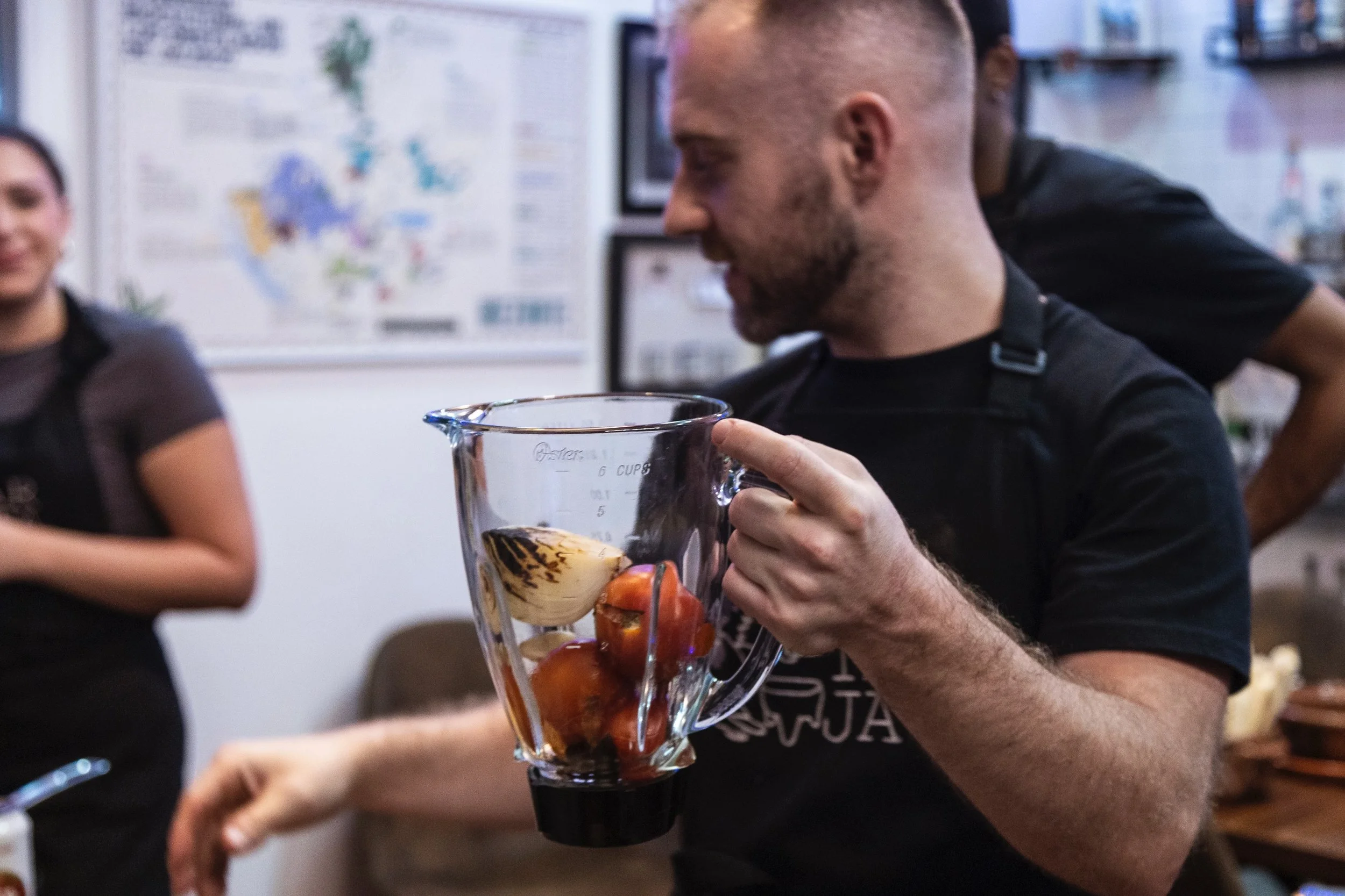 A person holding a glass pitcher filled with strawberries and ice, with a woman standing nearby, blurred background with a whiteboard and shelves.