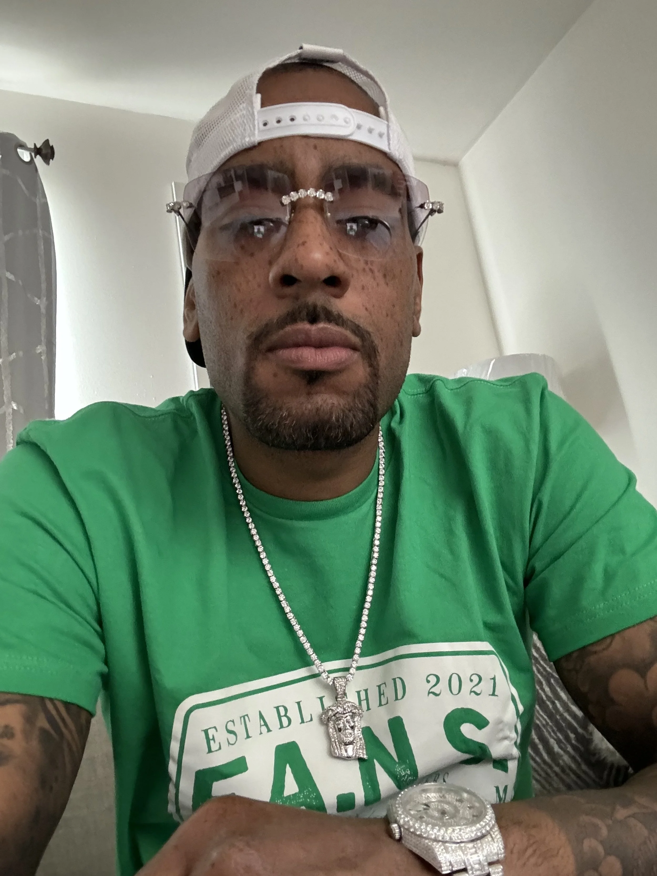 A man wearing a white cap backwards, clear eyeglasses with rhinestones, a green T-shirt with white lettering, a diamond chain, a large diamond watch, and a Jesus face pendant, sitting indoors.