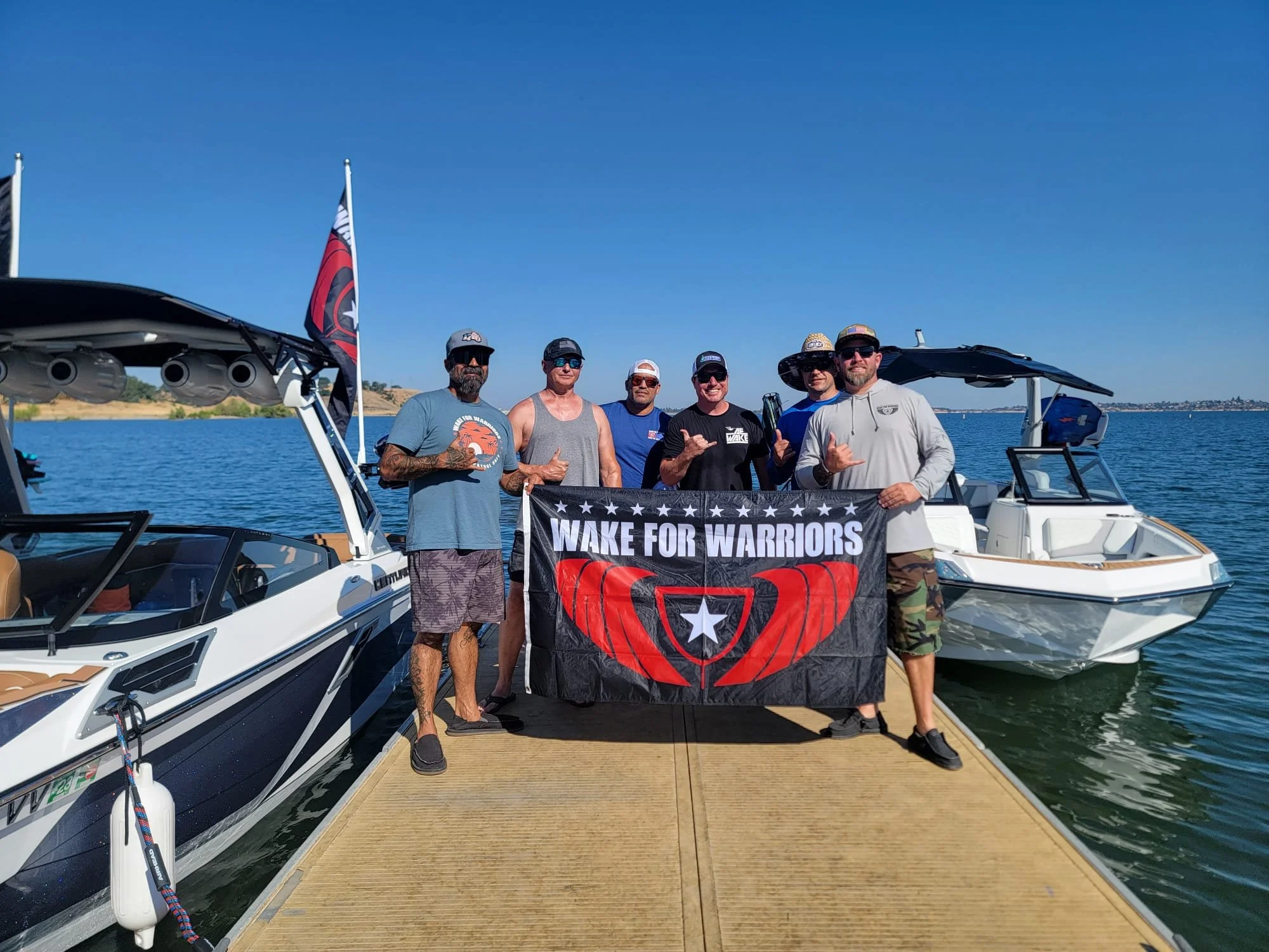 Group of six men on a dock holding a banner that reads 'Wake for Warriors' with boats and water in the background.