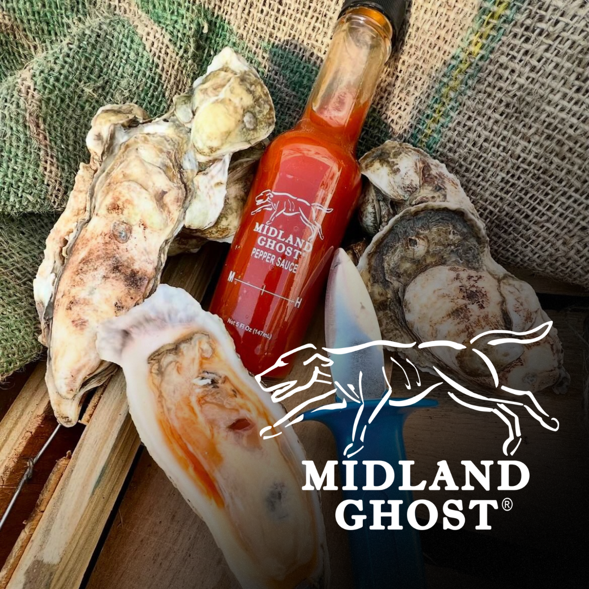 A bottle of Midland Ghost pepper sauce surrounded by oyster shells and a knife on a wooden surface, with burlap and green textured fabric in the background.