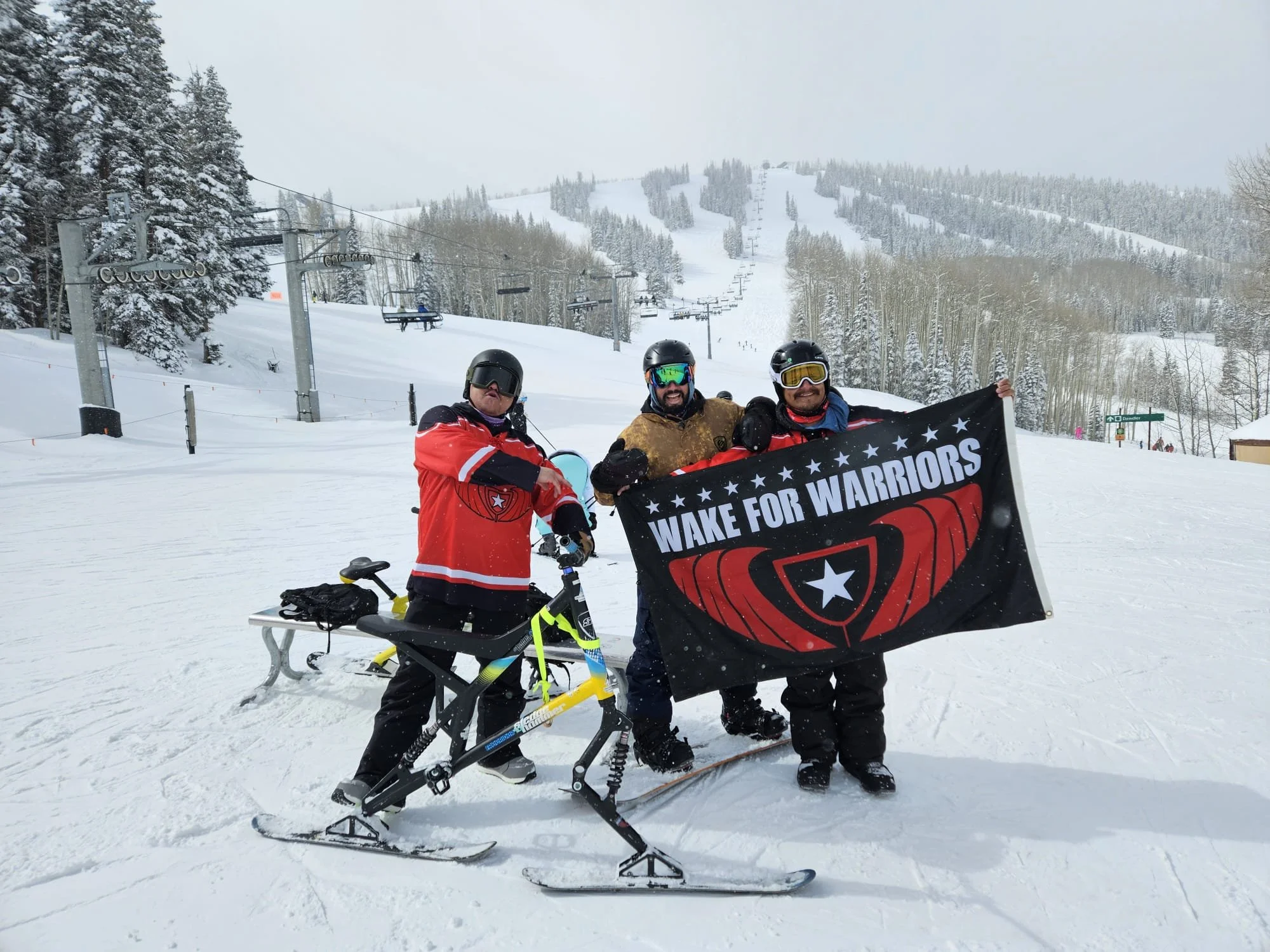 Three skiers standing on a snowy mountain holding a black flag with white and red design and text that reads "Wake for Warriors," with ski lifts and snow-covered trees in the background.