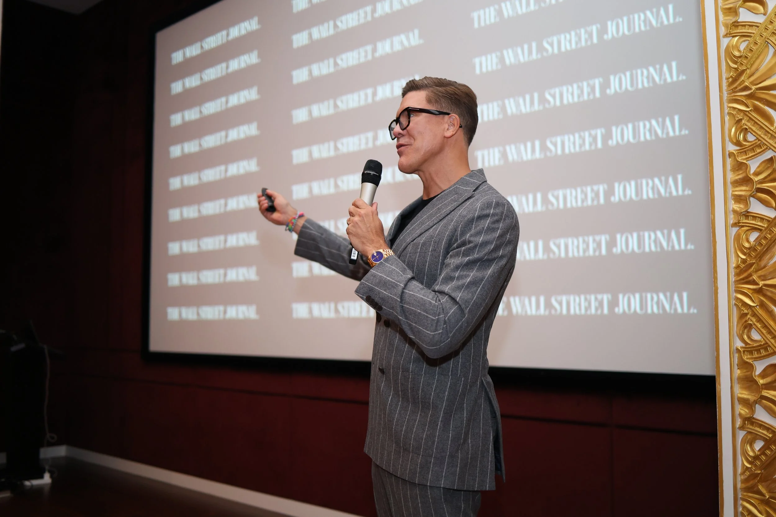 Man speaking into a microphone while seated on a stage, wearing a gray blazer, black shirt, black pants, and black sneakers. Background features colorful abstract art and a large screen.