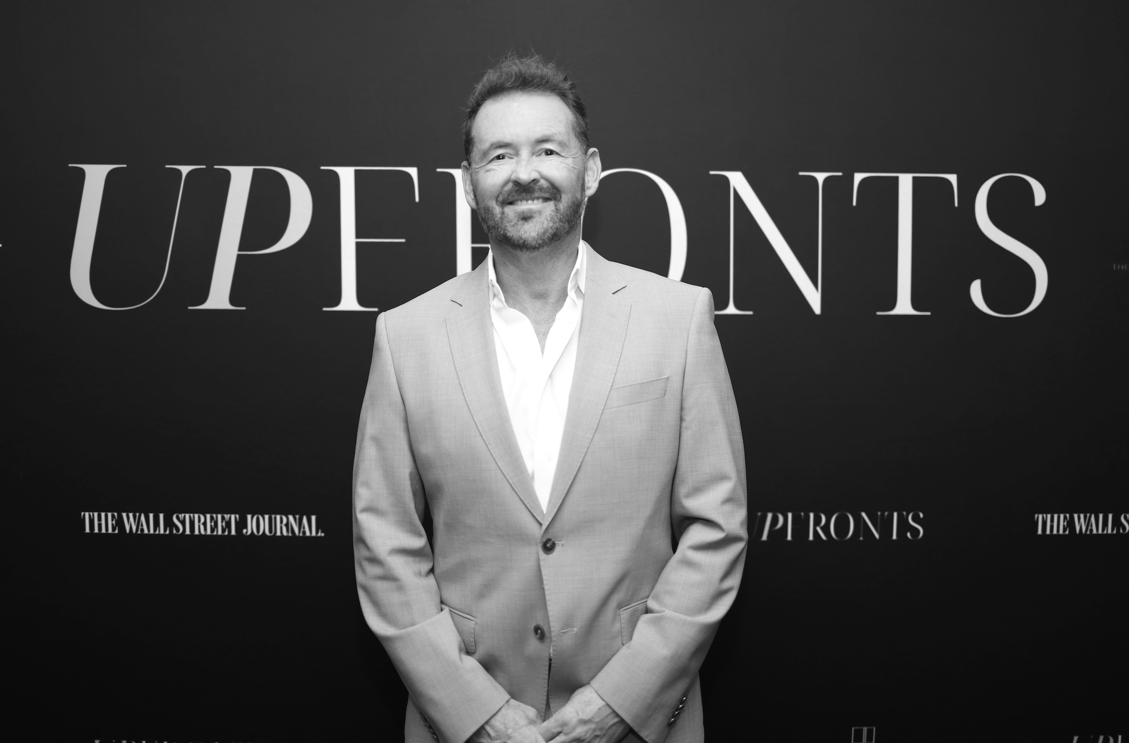 Black and white photo of a man in a suit standing indoors near a staircase, smiling.