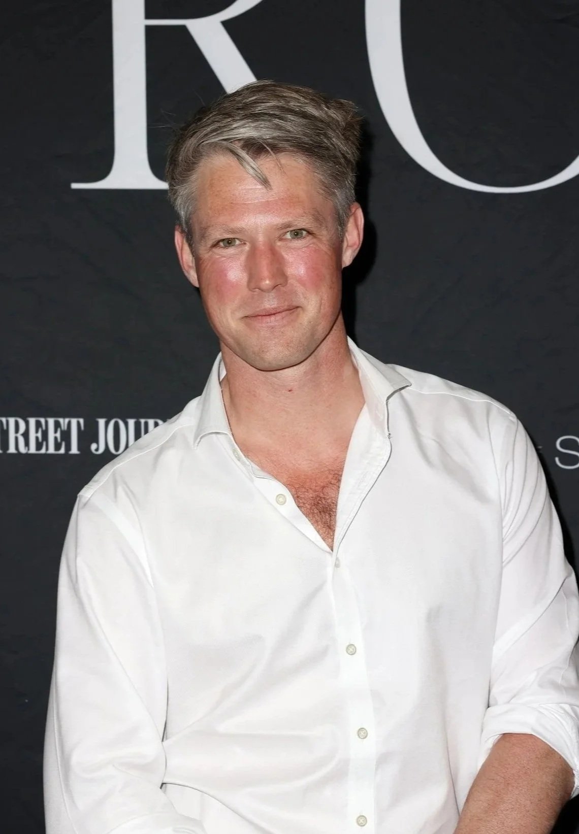 A man in a white shirt posing in front of a black backdrop with logos and text."