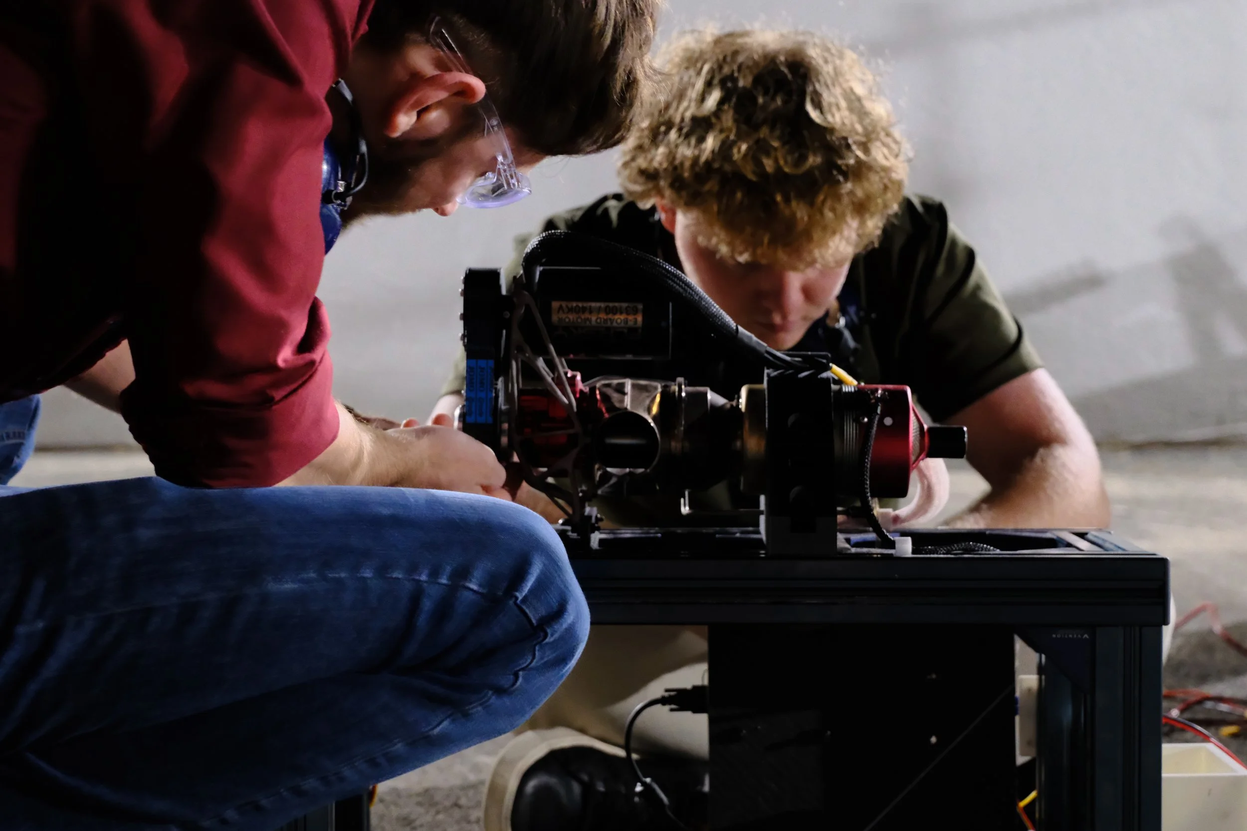 Two people working together on a mechanical or engineering project, focusing on a small machine or device on a black table, with a white wall in the background.