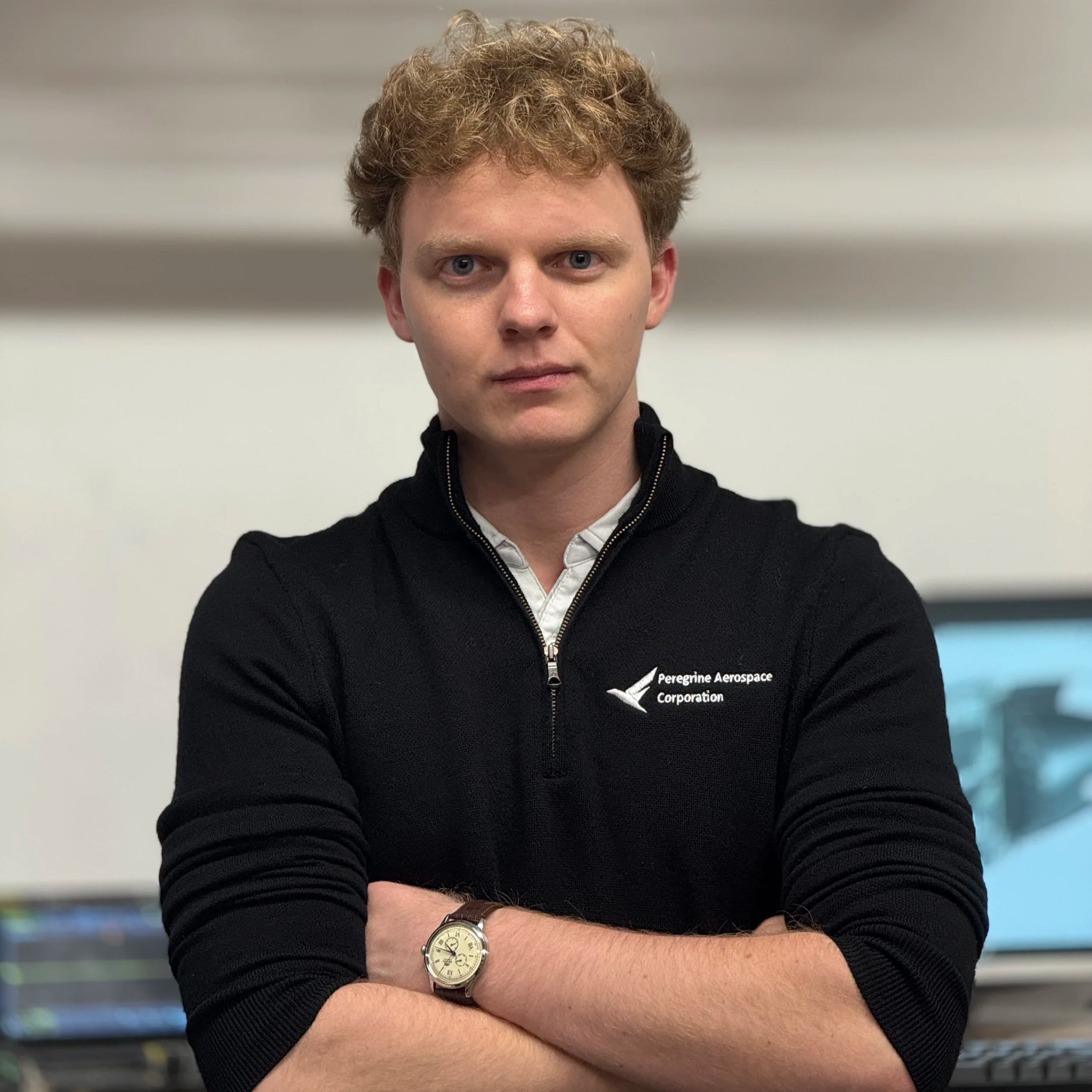 Donovan Shea, wearing a black quarter-zip sweater with the Peregrine Aerospace Corporation logo, standing with arms crossed in front of a computer screen in an workshop environment.
