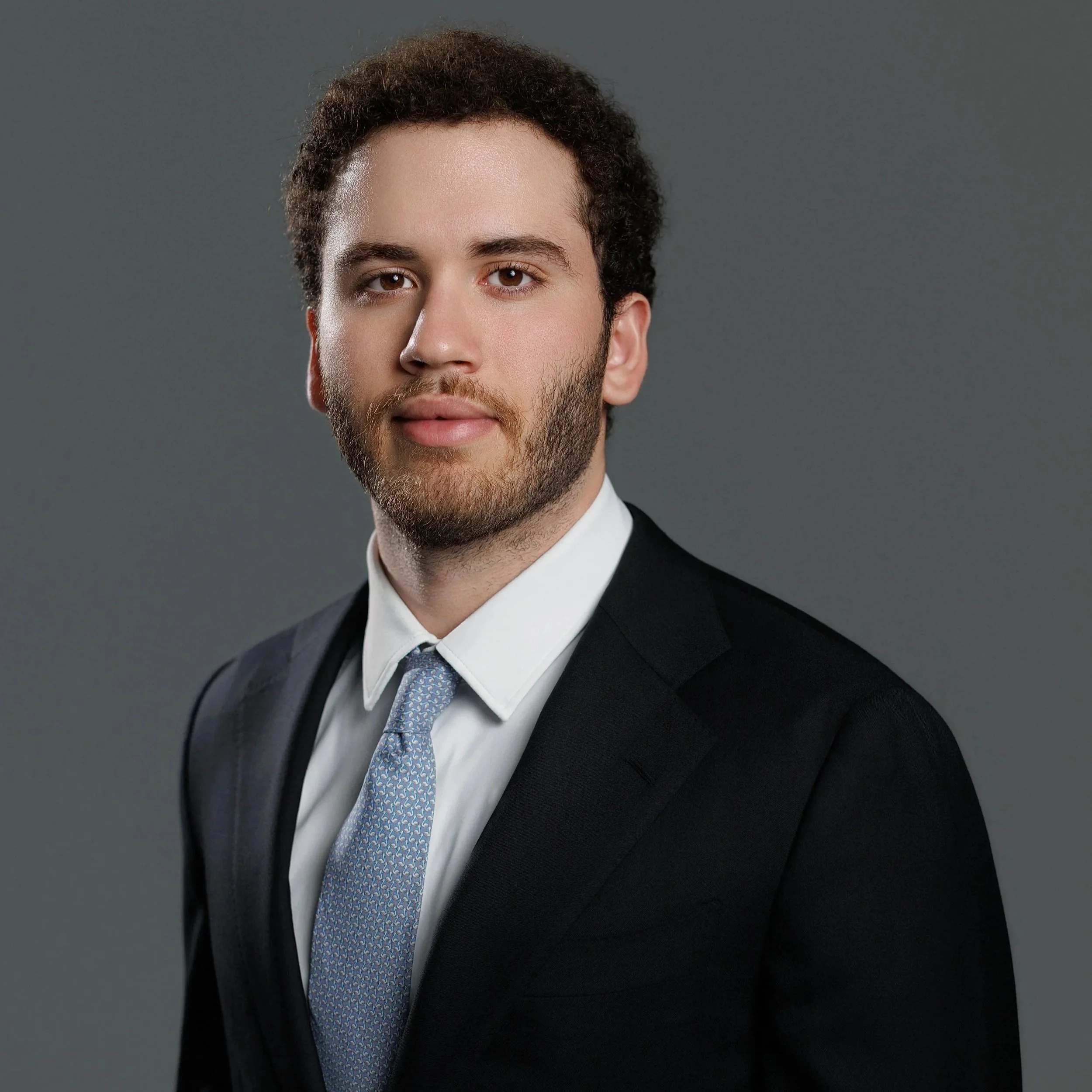 A man with glasses and a beard, wearing a white shirt and dark blazer, standing indoors near a metallic structure, possibly a piece of equipment or furniture.
