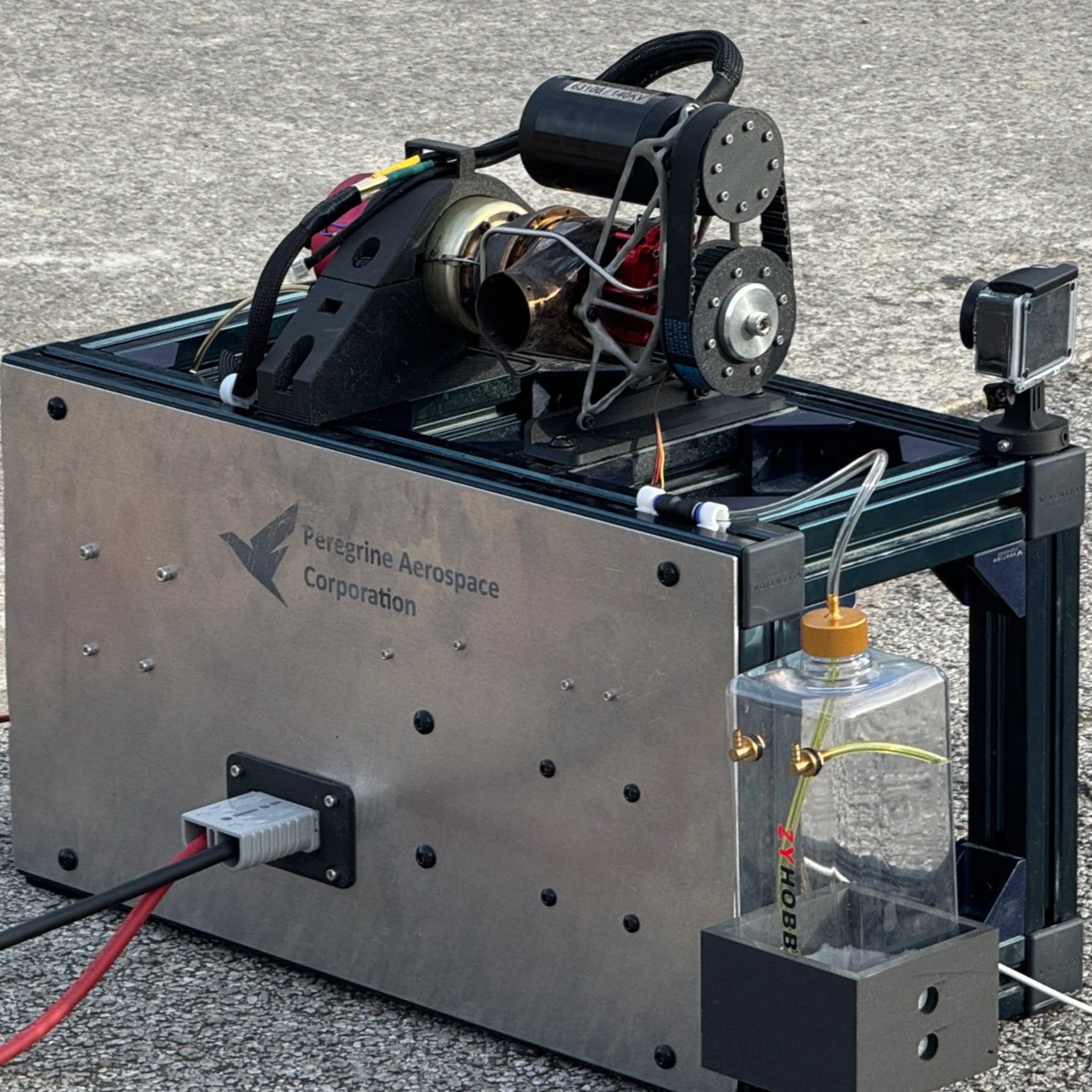 Close-up of a jet engine or turbine with black and gold components, wires, and a cylindrical motor labeled 'FLYSKY.' The device is mounted on a black frame with visible wires and electronic components below.