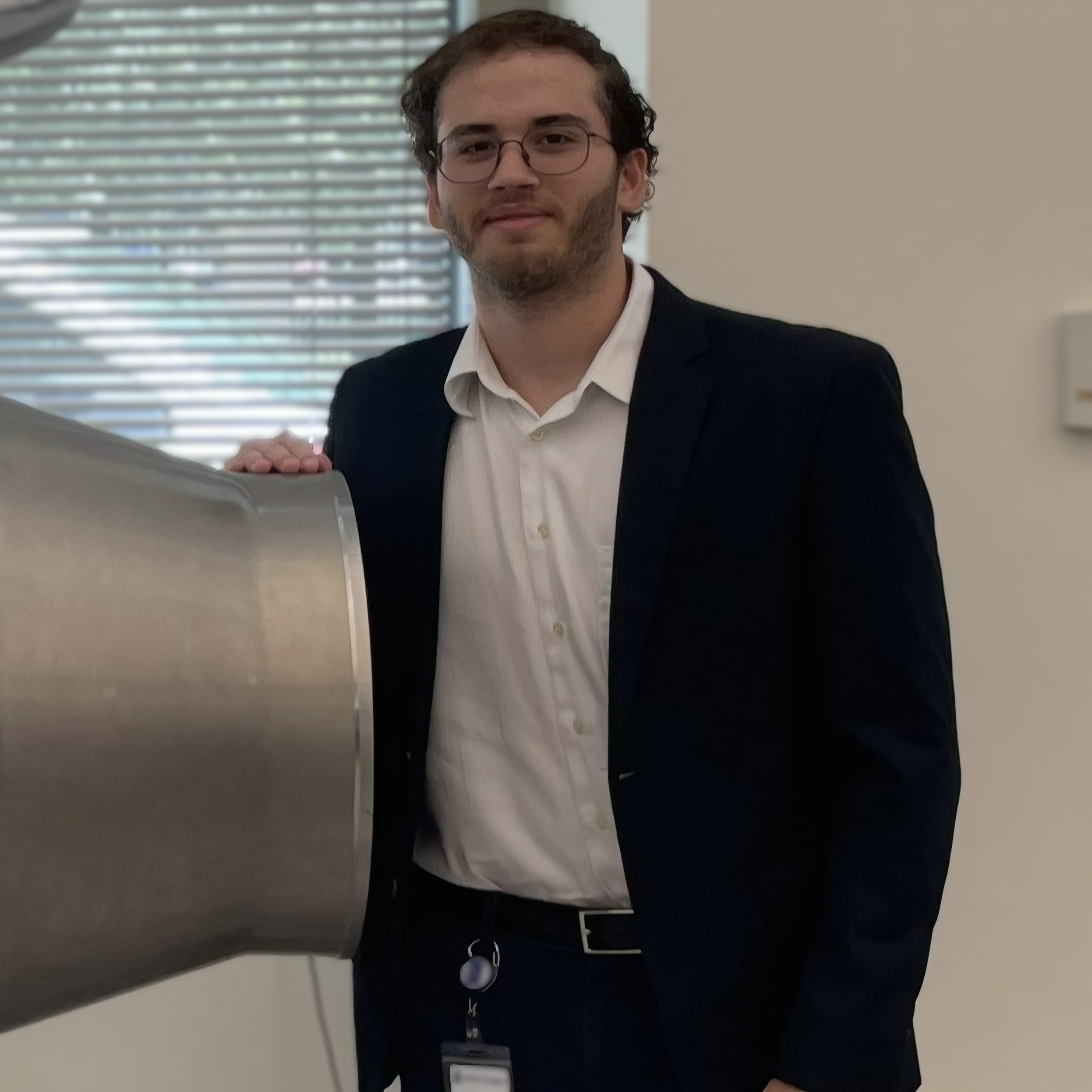 A man with glasses and a beard, wearing a white shirt and dark blazer, standing indoors near a metallic structure, possibly a piece of equipment or furniture.