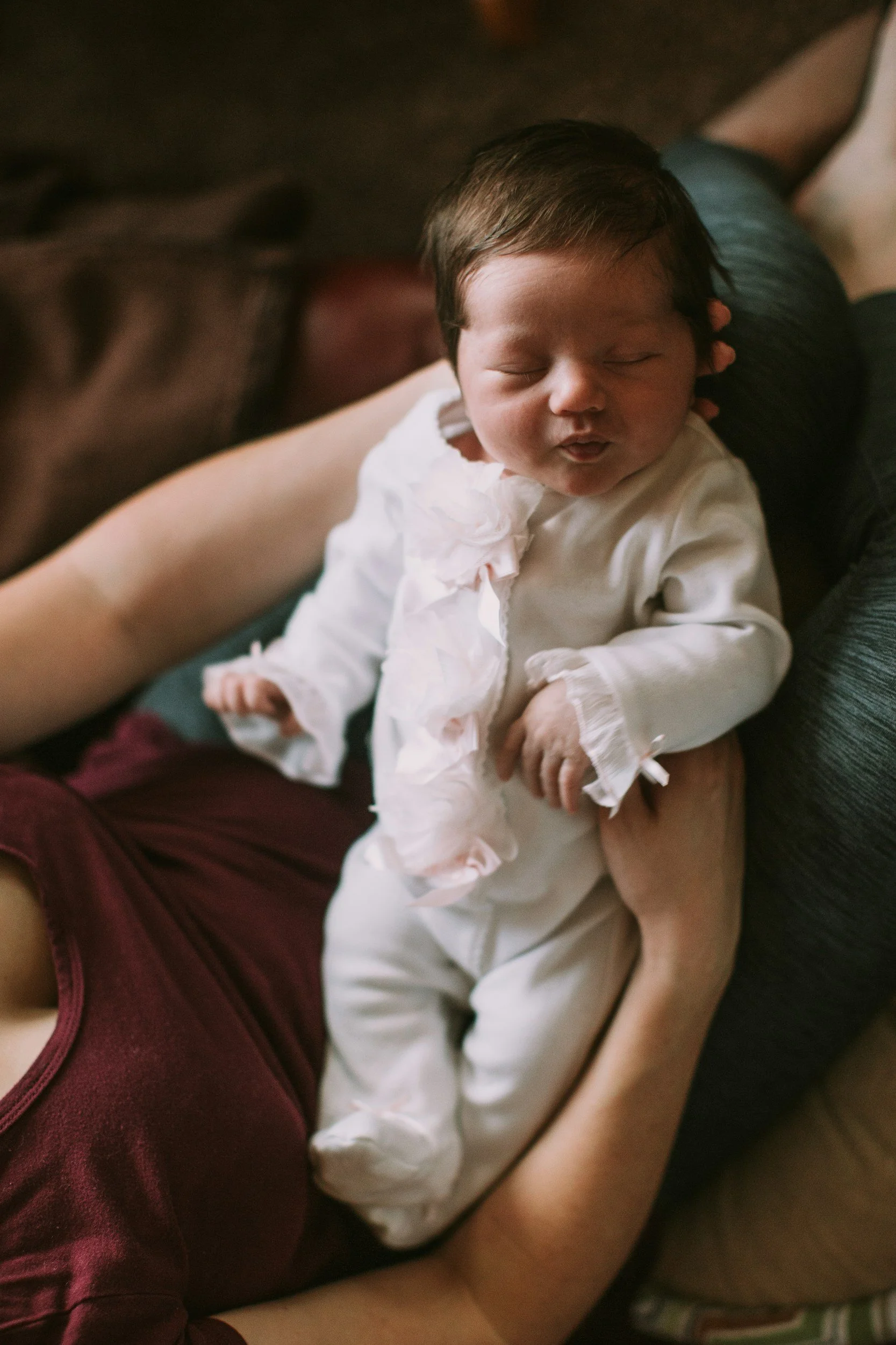 A newborn baby with dark hair, dressed in a white outfit with lace details, lying on a person's chest, with their eyes closed and lips puckered as if making a fish face.