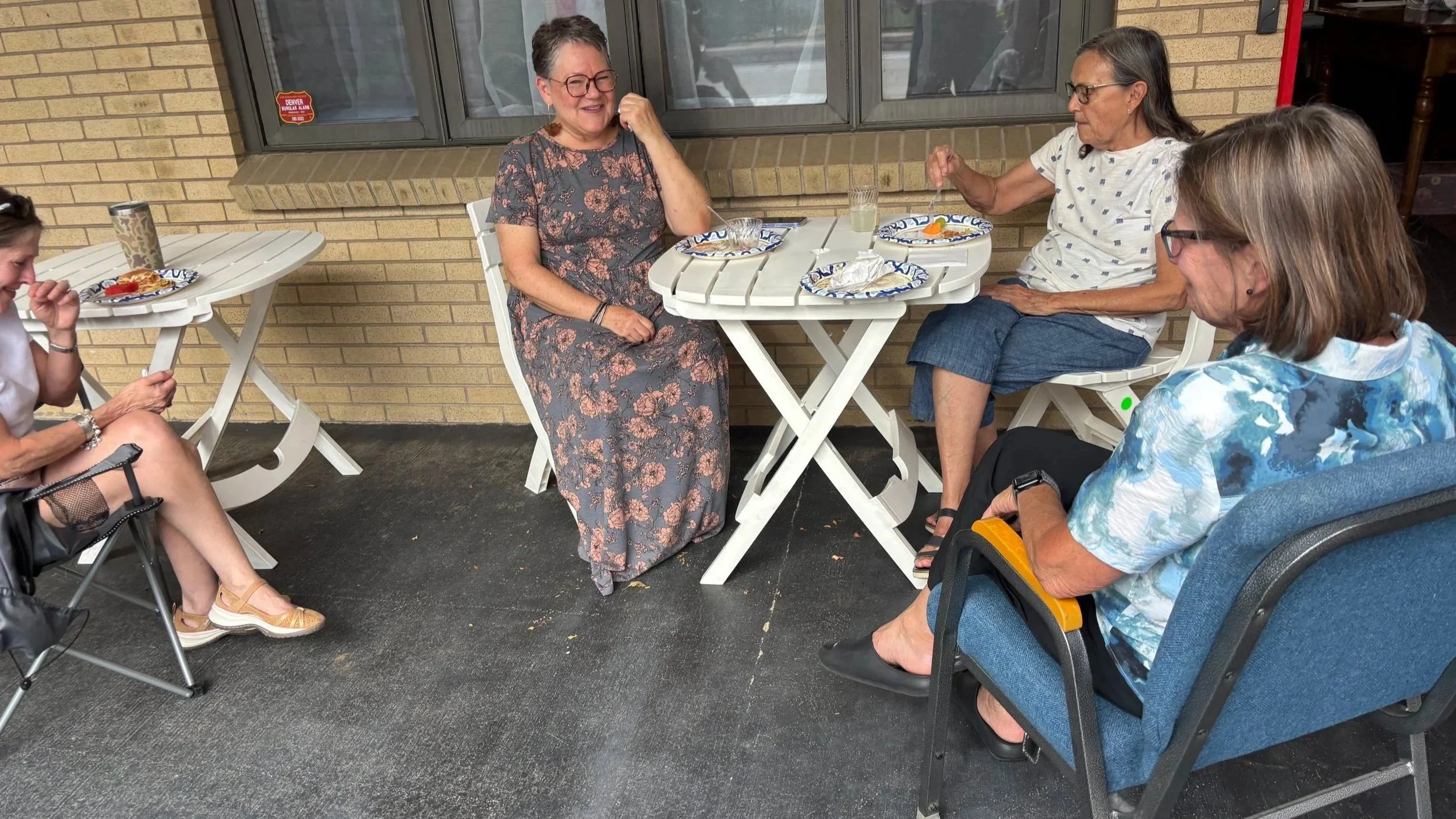 Four women sitting outside at a table, enjoying a meal and drinks, engaging in conversation and smiling.