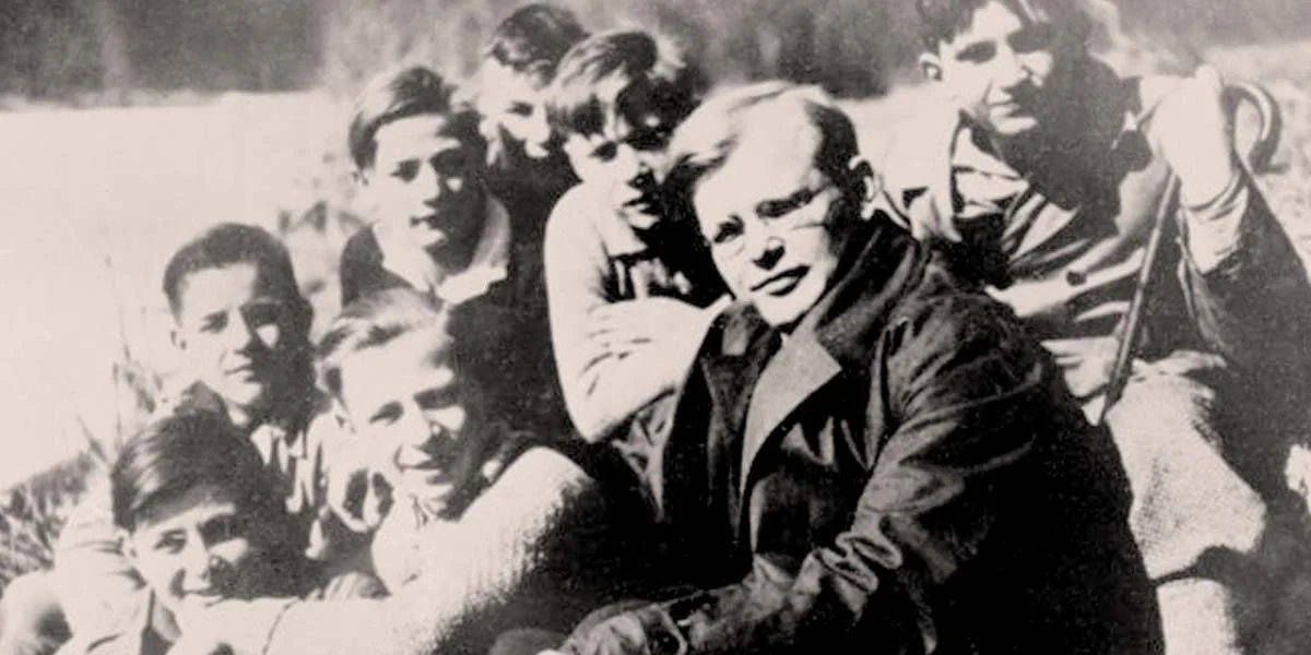 Bonhoeffer in a coat looks at the camera, boys are piled up behind to pose with him