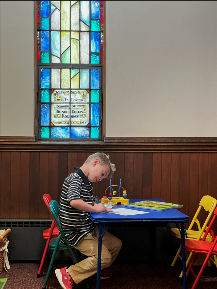 A young boy playing with toys at a small blue table in a room with stained glass window and wood paneling.