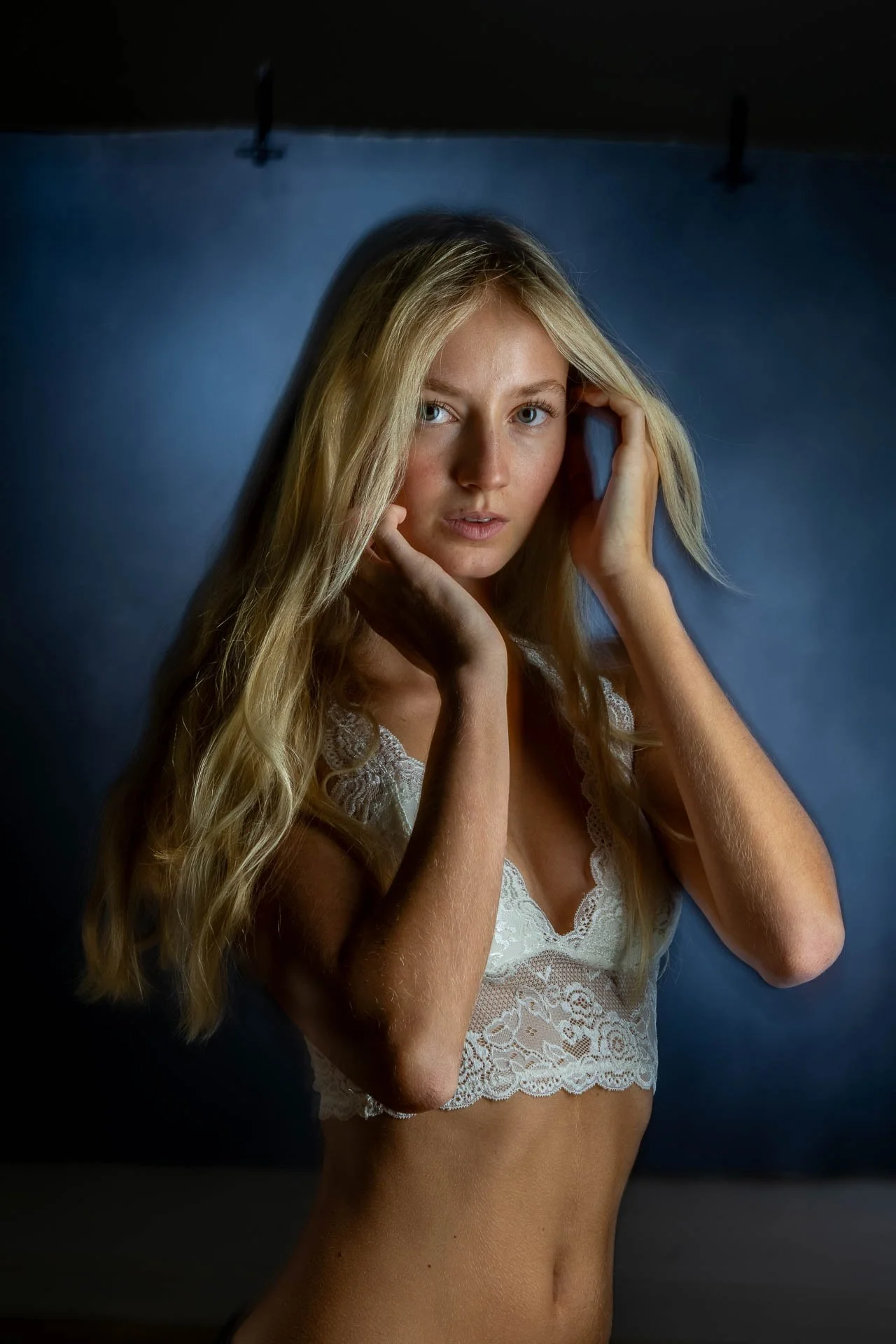 Porträt einer jungen Frau mit langen, lockigen blonden Haaren, blauen Augen und heller Haut, die entspannt die Hände im Haar hat, vor einem blauen Hintergrund.