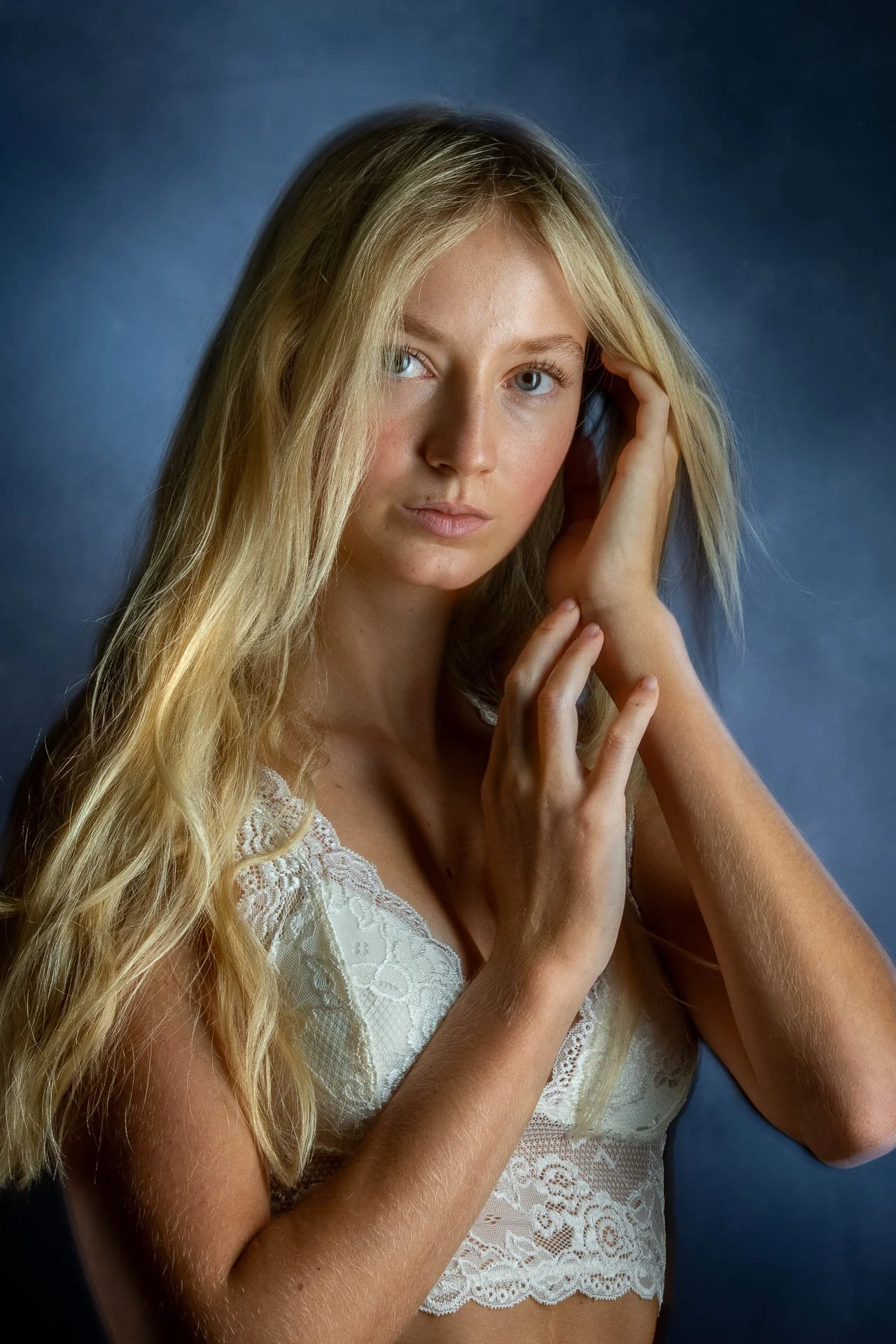Porträt eines jungen Mädchens mit langen blonden Haaren, blauen Augen und hellem Hauttyp, trägt eine weiße Spitze-Top, hält eine Hand an ihr Gesicht, steht vor einem blauen Hintergrund.