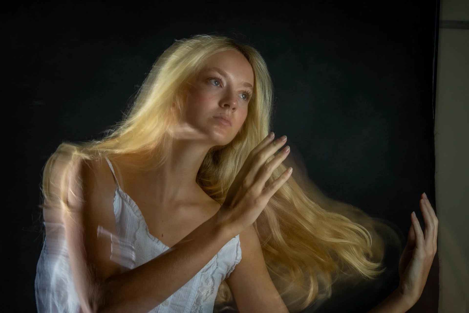 Porträt einer blonden Frau in weißem Kleid, die in eine Kamera schaut, vor dunklem Hintergrund.