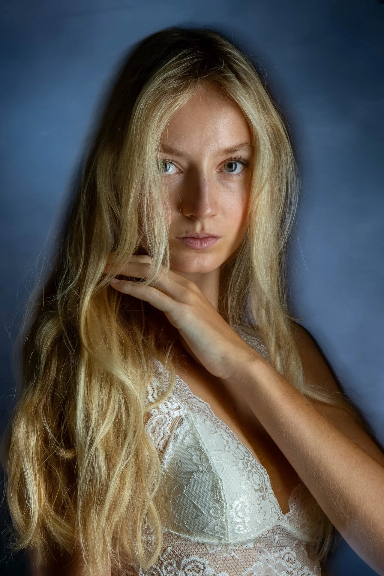 Nahaufnahme eines jungen Mädchens mit langen, lockigen, blonden Haaren und blauen Augen vor einem blauen Hintergrund, das eine Hand an den Hals legt.